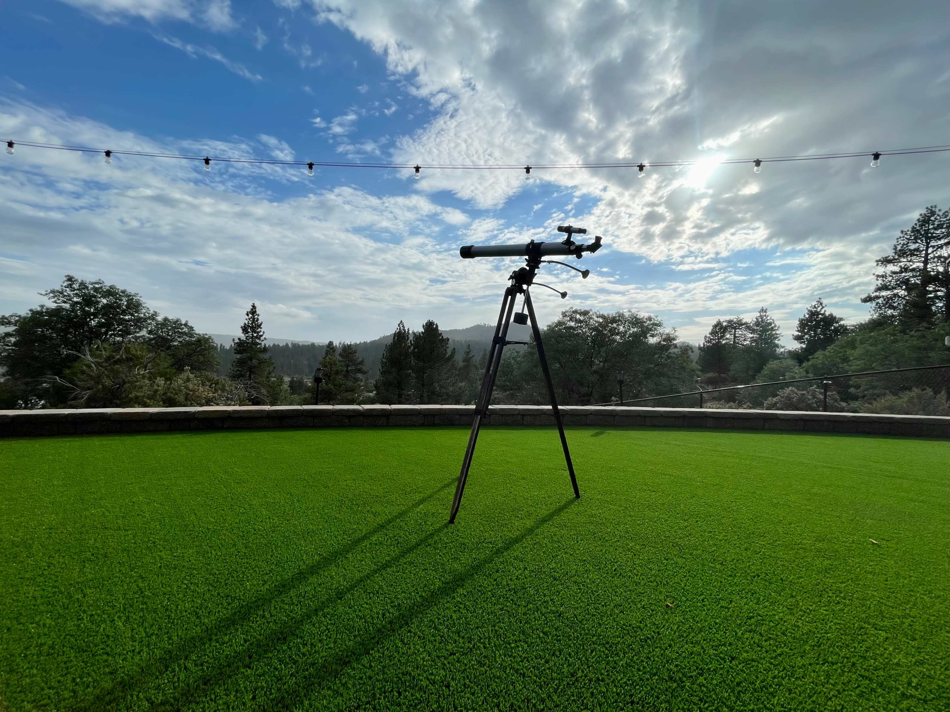 A telescope is set up on a grassy area with trees and clouds in the background.