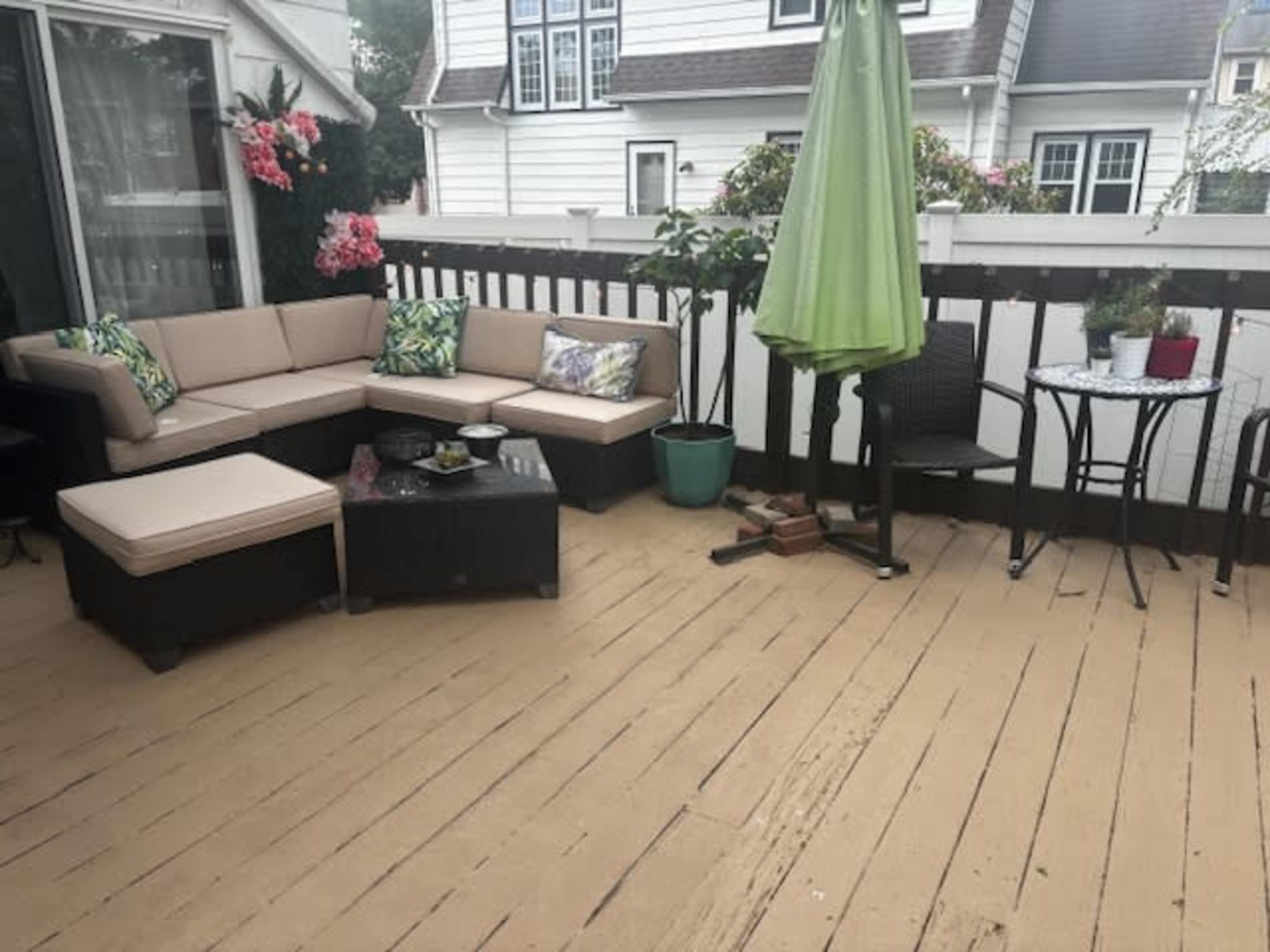 The image shows a deck area with a sectional sofa, a small table with chairs, and potted plants surrounded by a wooden railing.