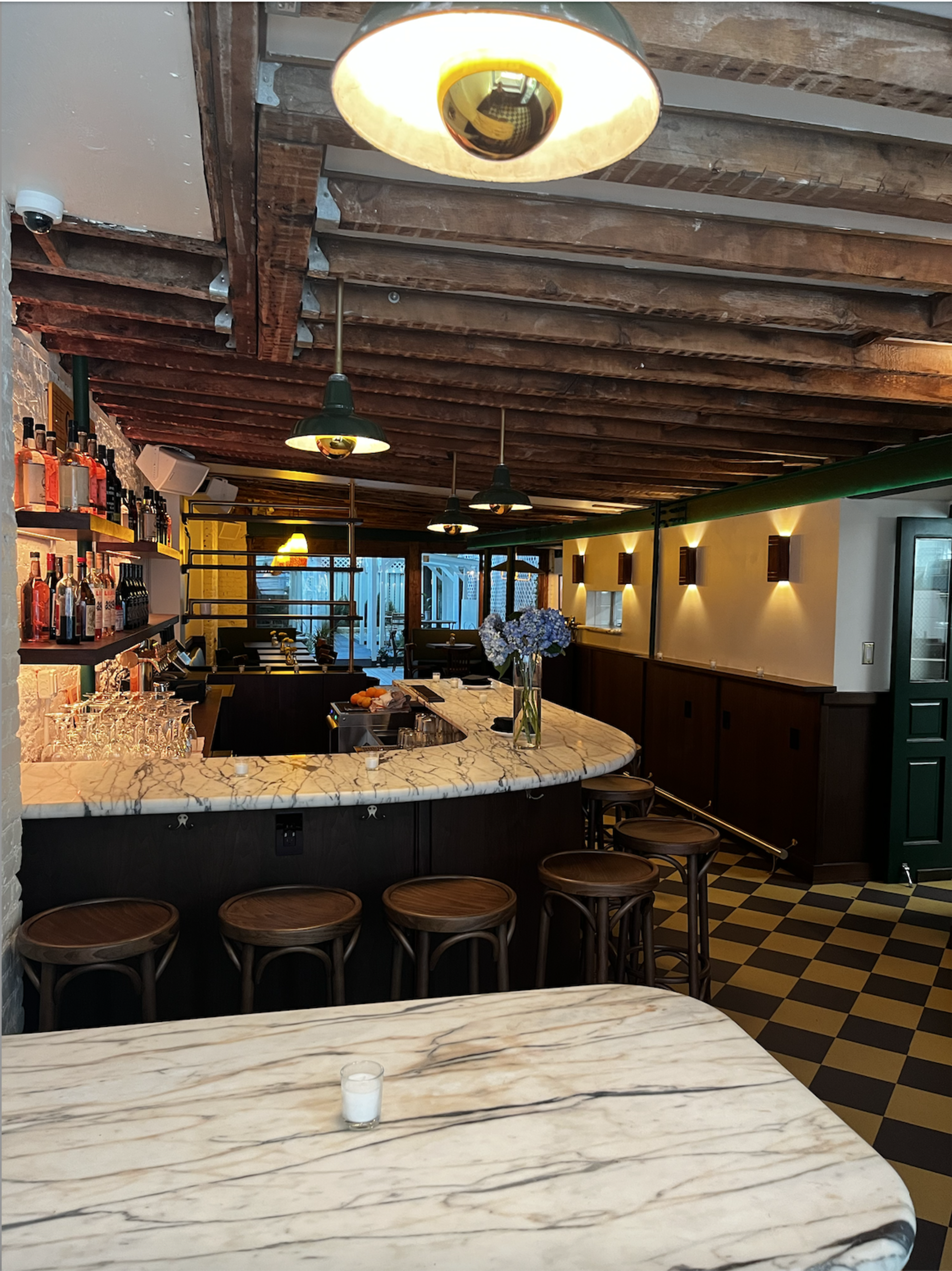 The image shows a bar area with a marble countertop, wooden stools, and exposed ceiling beams, featuring shelves of bottles in the background.