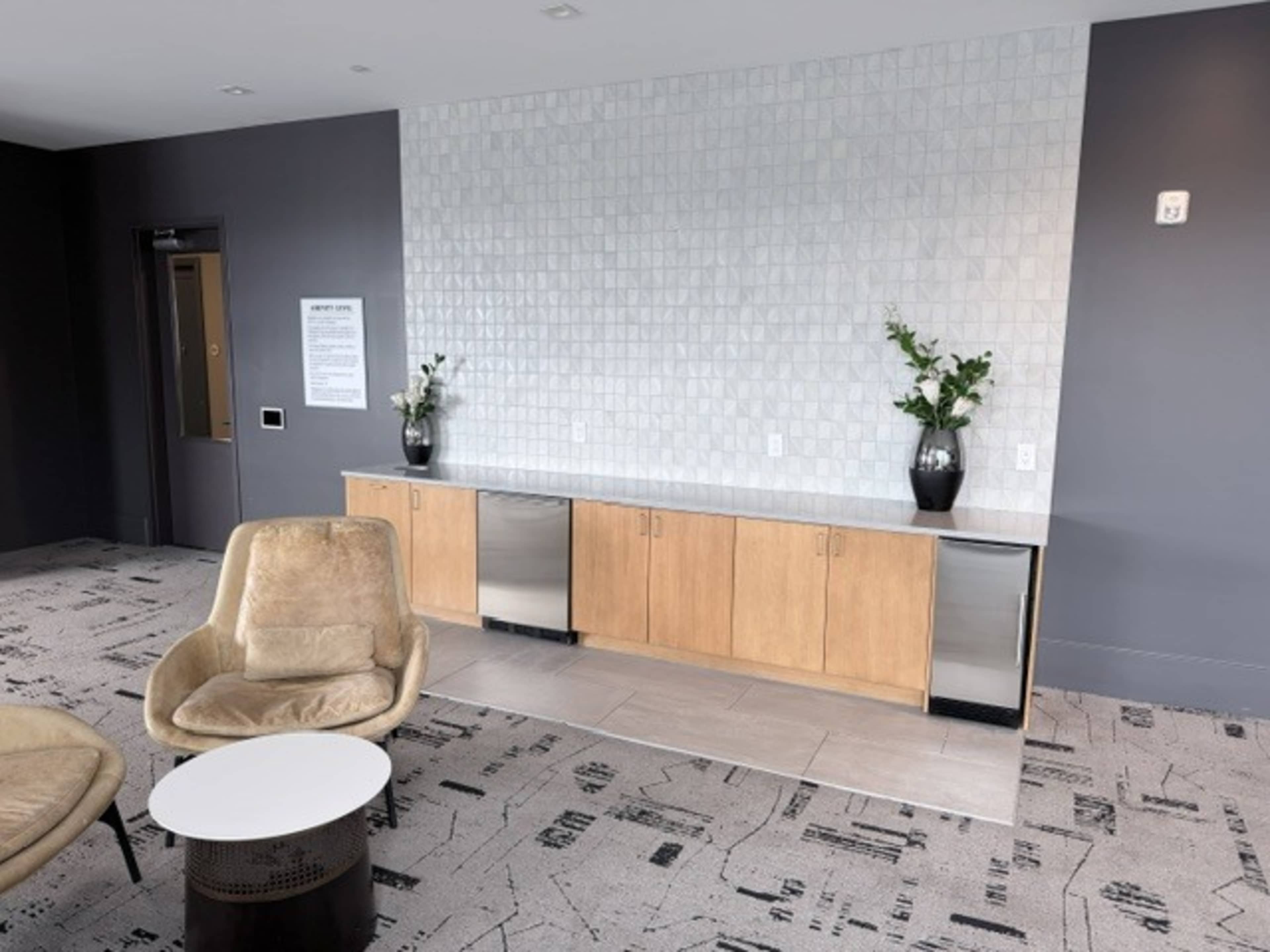 The image shows a modern lounge area featuring a light-colored cabinet with a countertop, an armchair, a round table, and decorative plants on display.