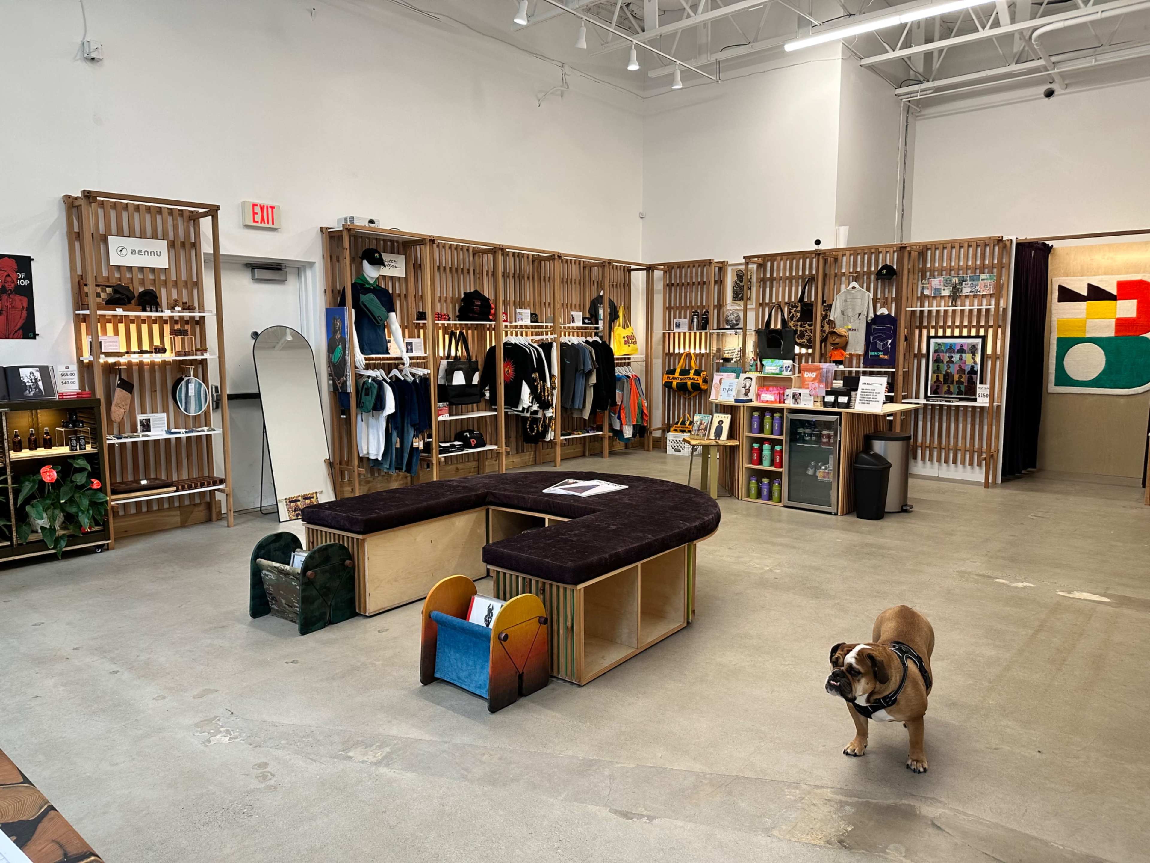 A retail store with wooden shelving displaying clothing and merchandise, a central seating area, and a dog on the floor.