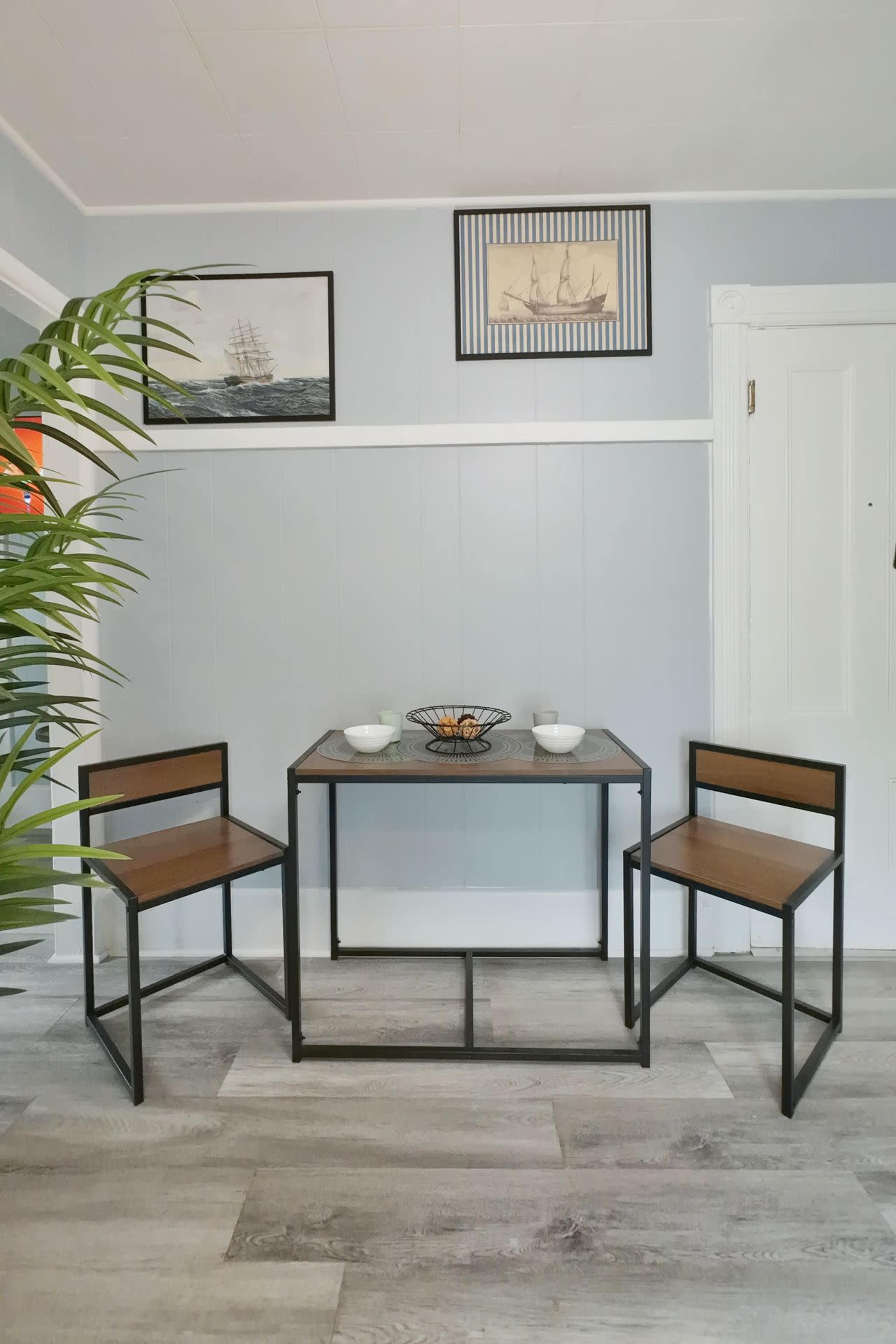 A small dining area features a black metal table with two matching chairs, accompanied by decorative bowls, and wall art depicting ships.