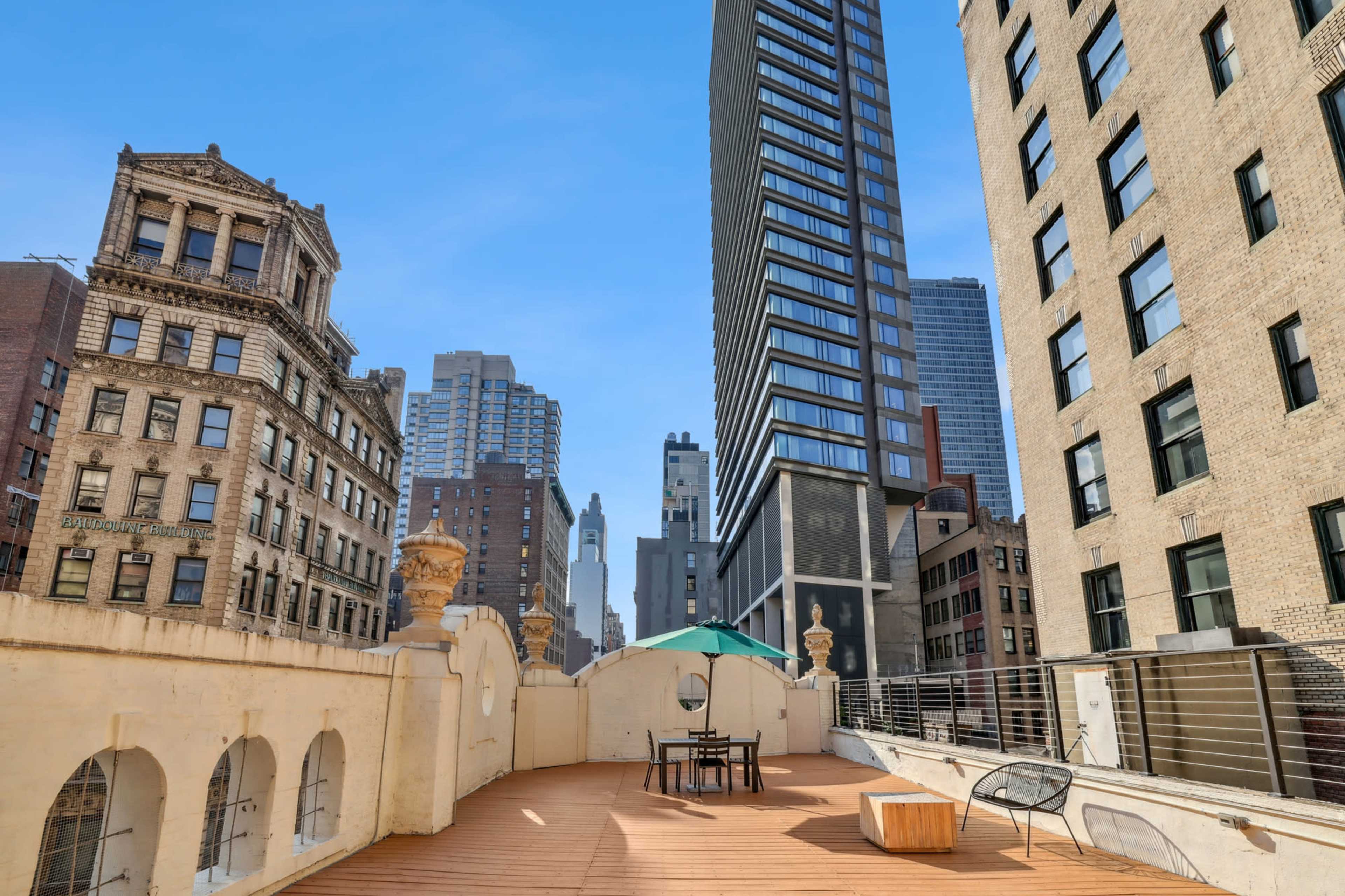 A rooftop terrace features a wooden deck with a table and chairs, surrounded by tall buildings in an urban setting.
