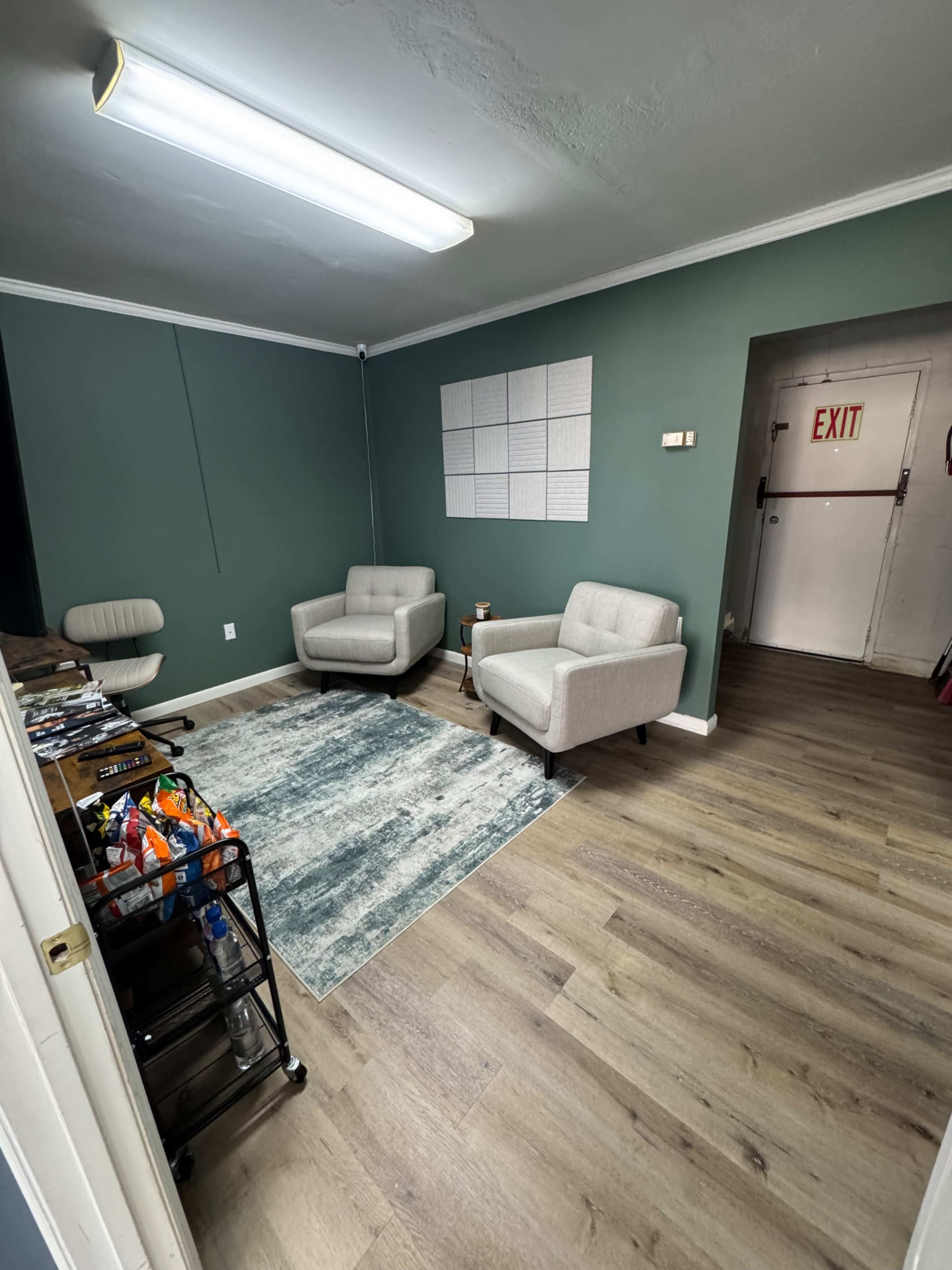 The image shows a small, vacant room with two gray chairs, a light blue area rug, a wall with a grid pattern, and an exit door on one side.