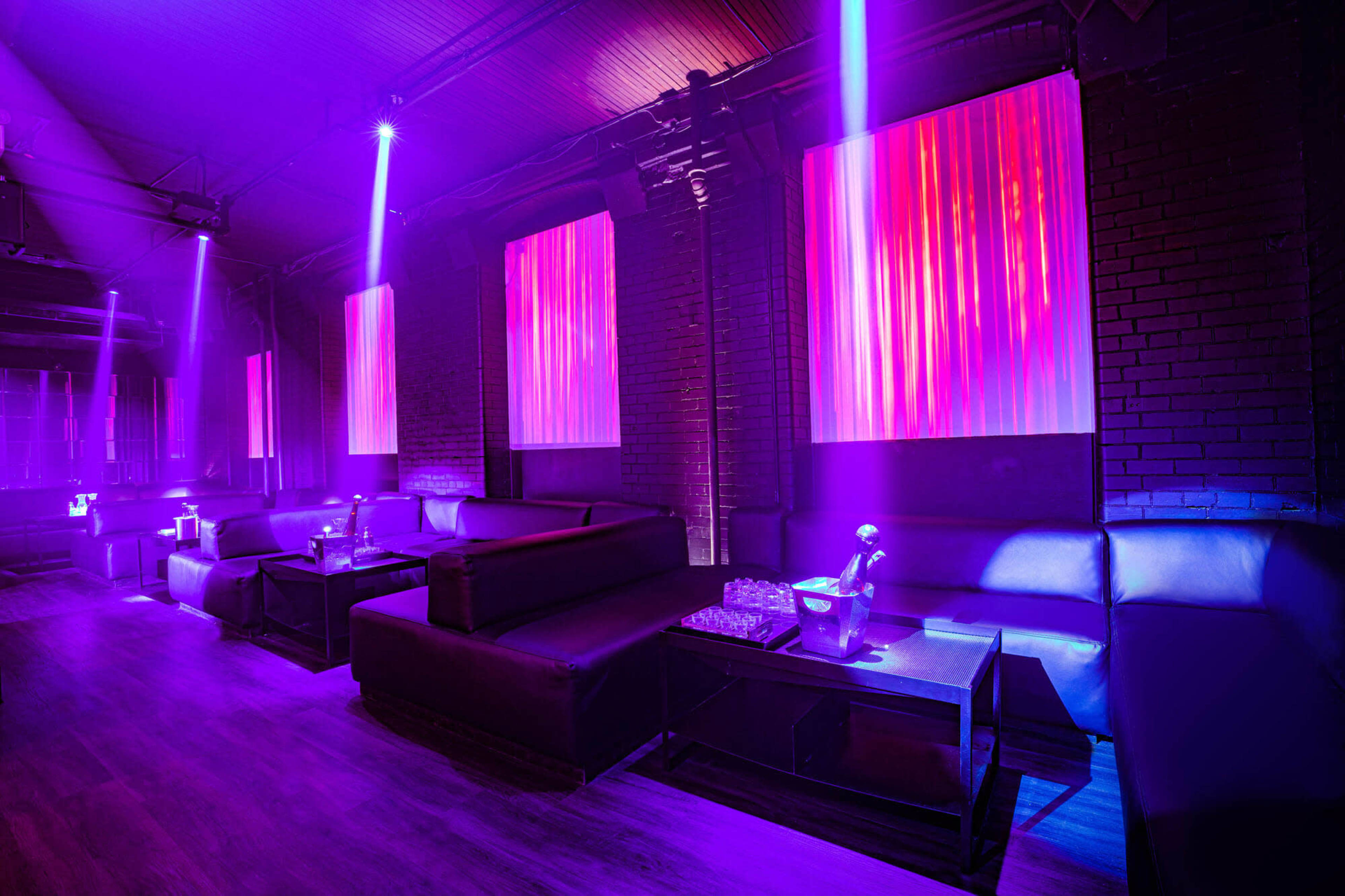 The image shows a dimly lit lounge with dark furniture, illuminated by purple lighting and featuring large screens displaying colorful patterns.