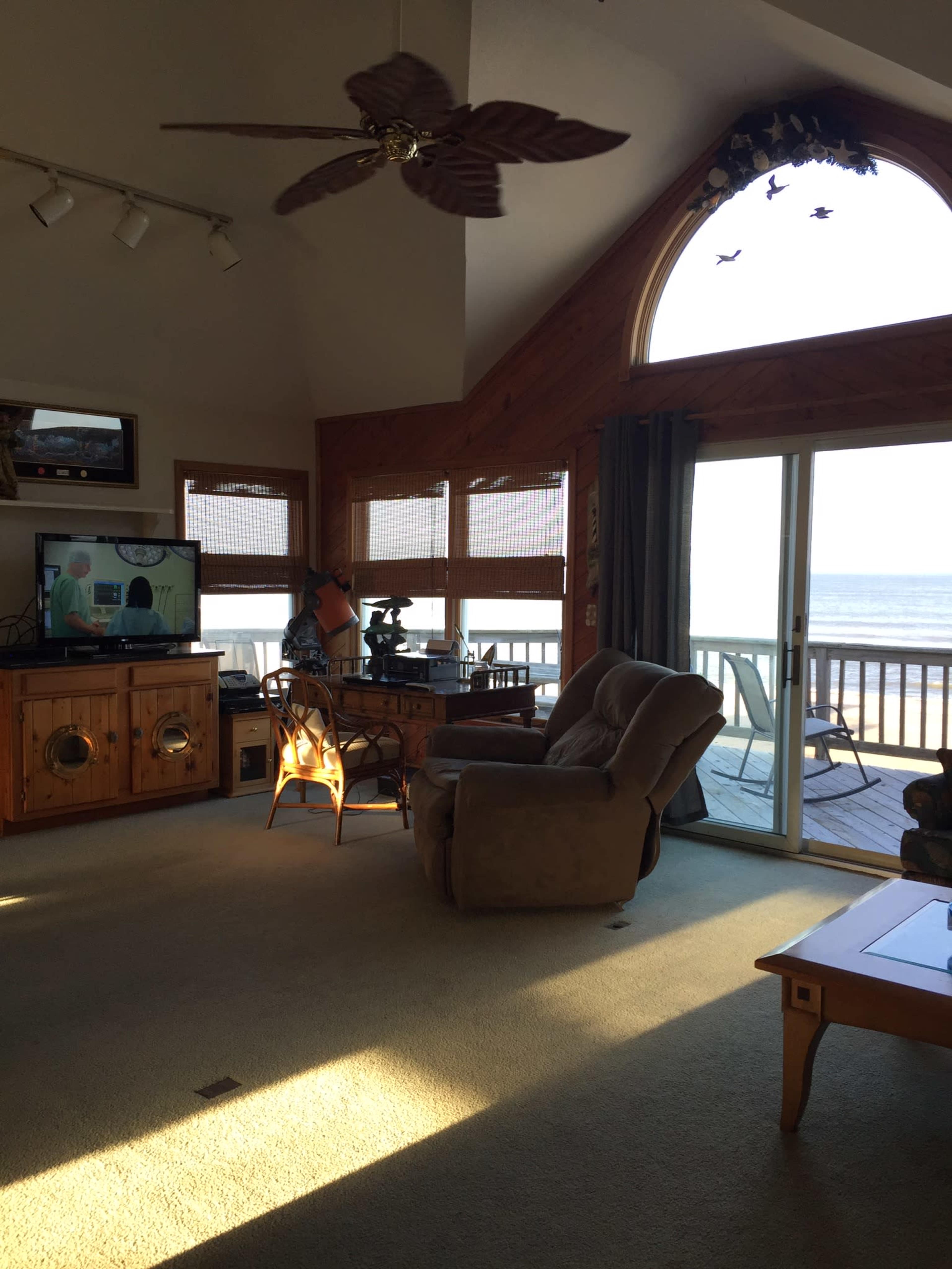 A light-filled living room features a comfortable seating area, a television, and a large window overlooking a deck by the water.