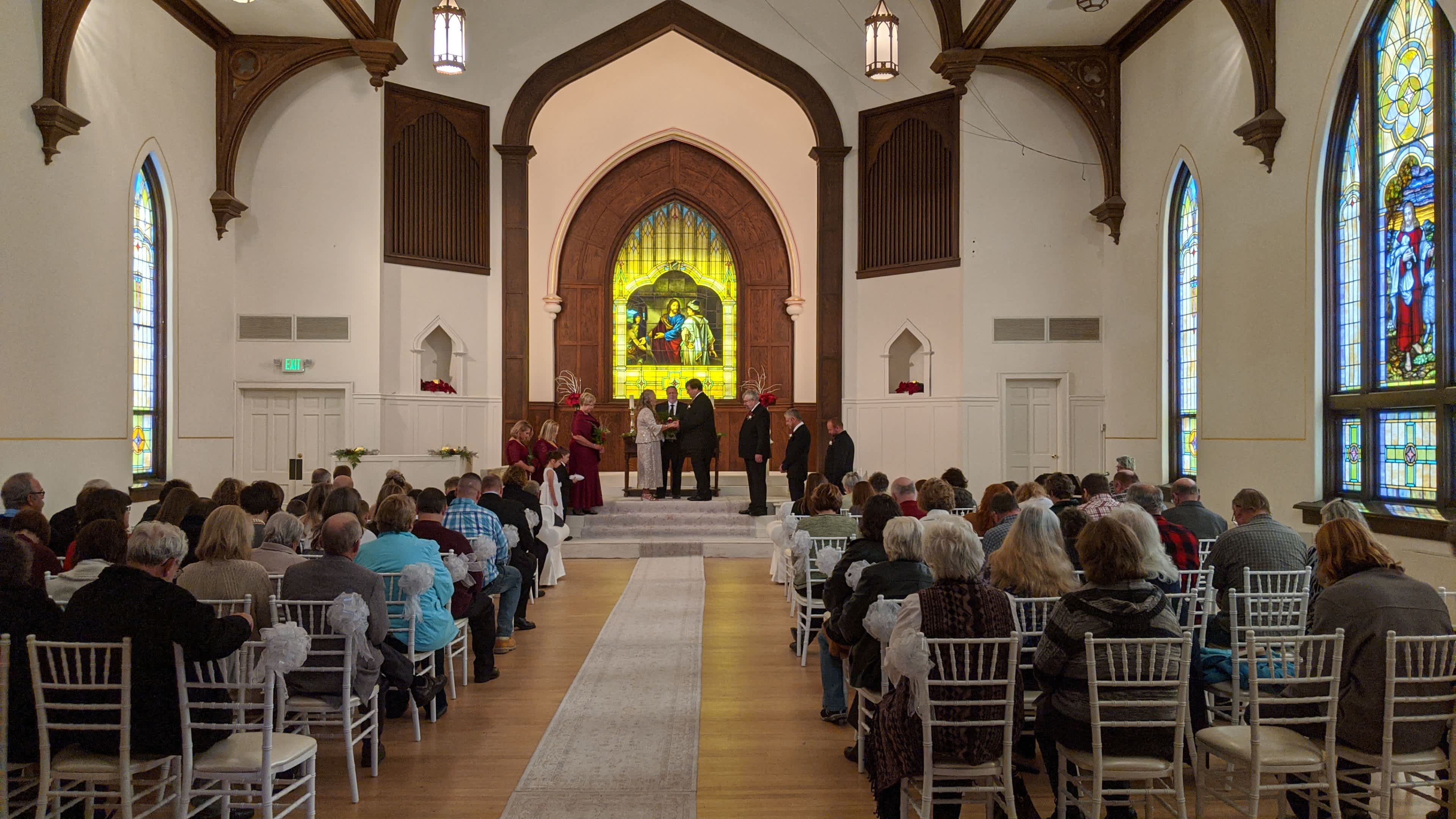 A wedding ceremony is taking place in a church, with a group of people gathered at the front and attendees seated in rows facing them.