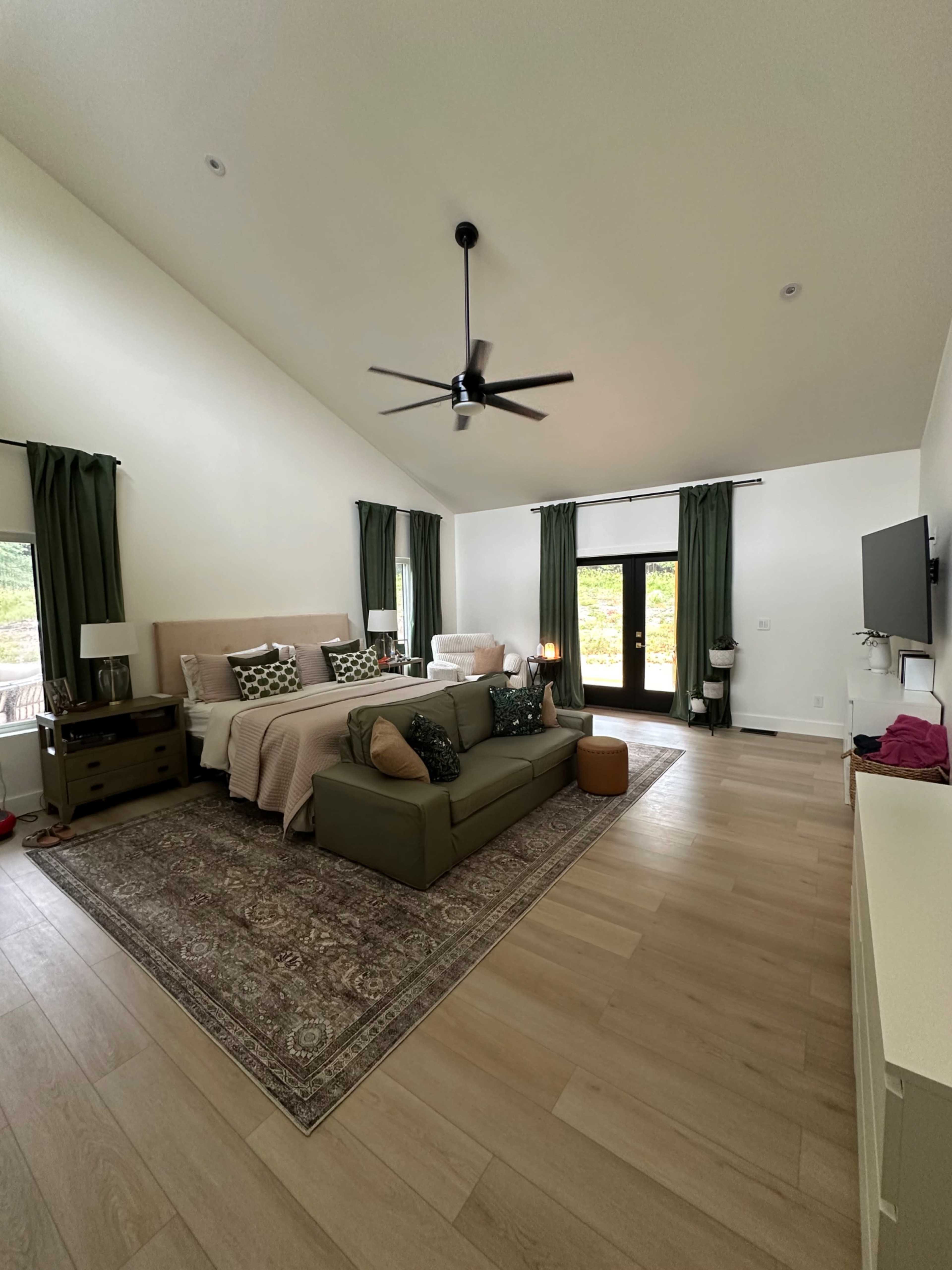 The image shows a spacious bedroom furnished with a bed, a sofa, and large windows with greenery visible outside.