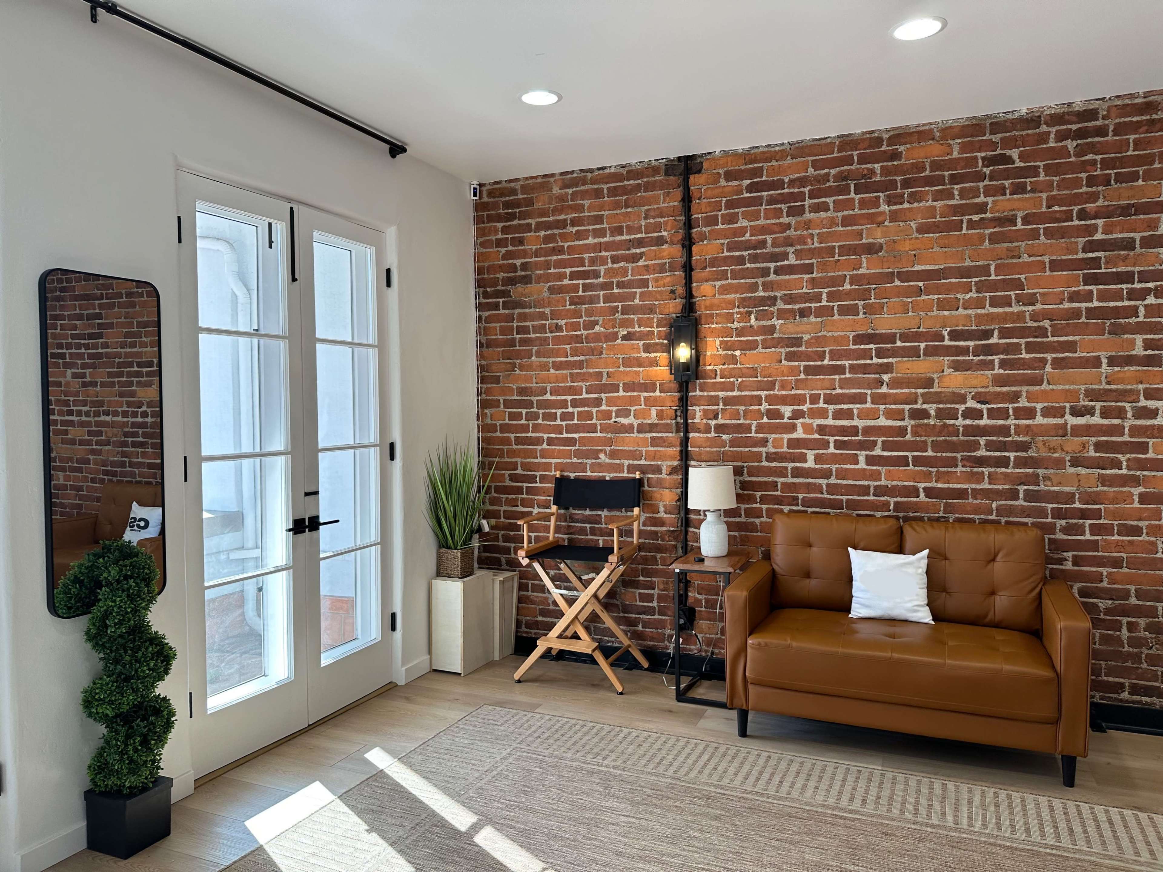 The image shows a cozy interior featuring a brown leather sofa, a director's chair, a small plant, and a brick wall with large glass doors.