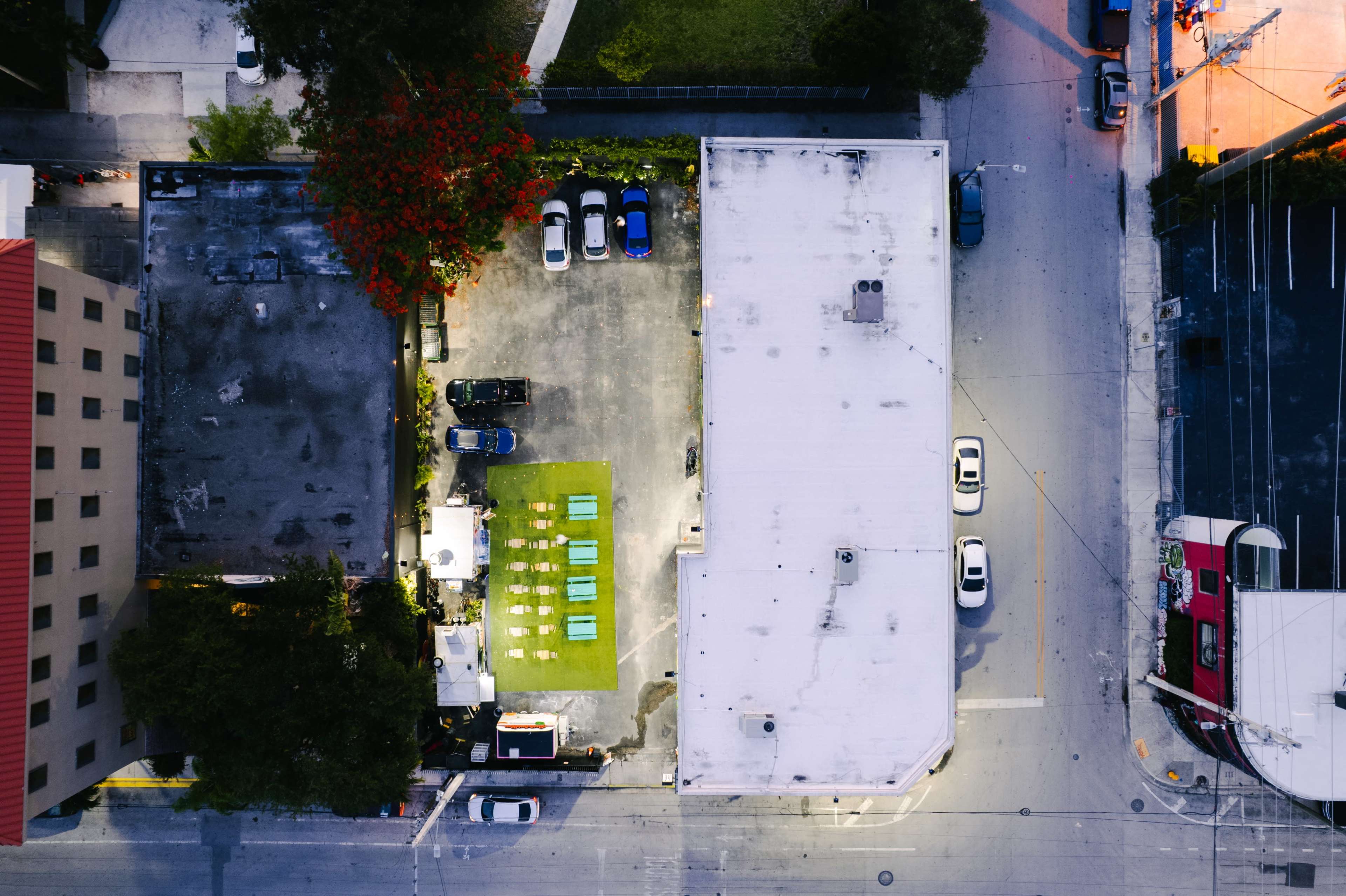 An aerial view shows a parking lot with several cars and a green area featuring lounge chairs, surrounded by buildings and streets.