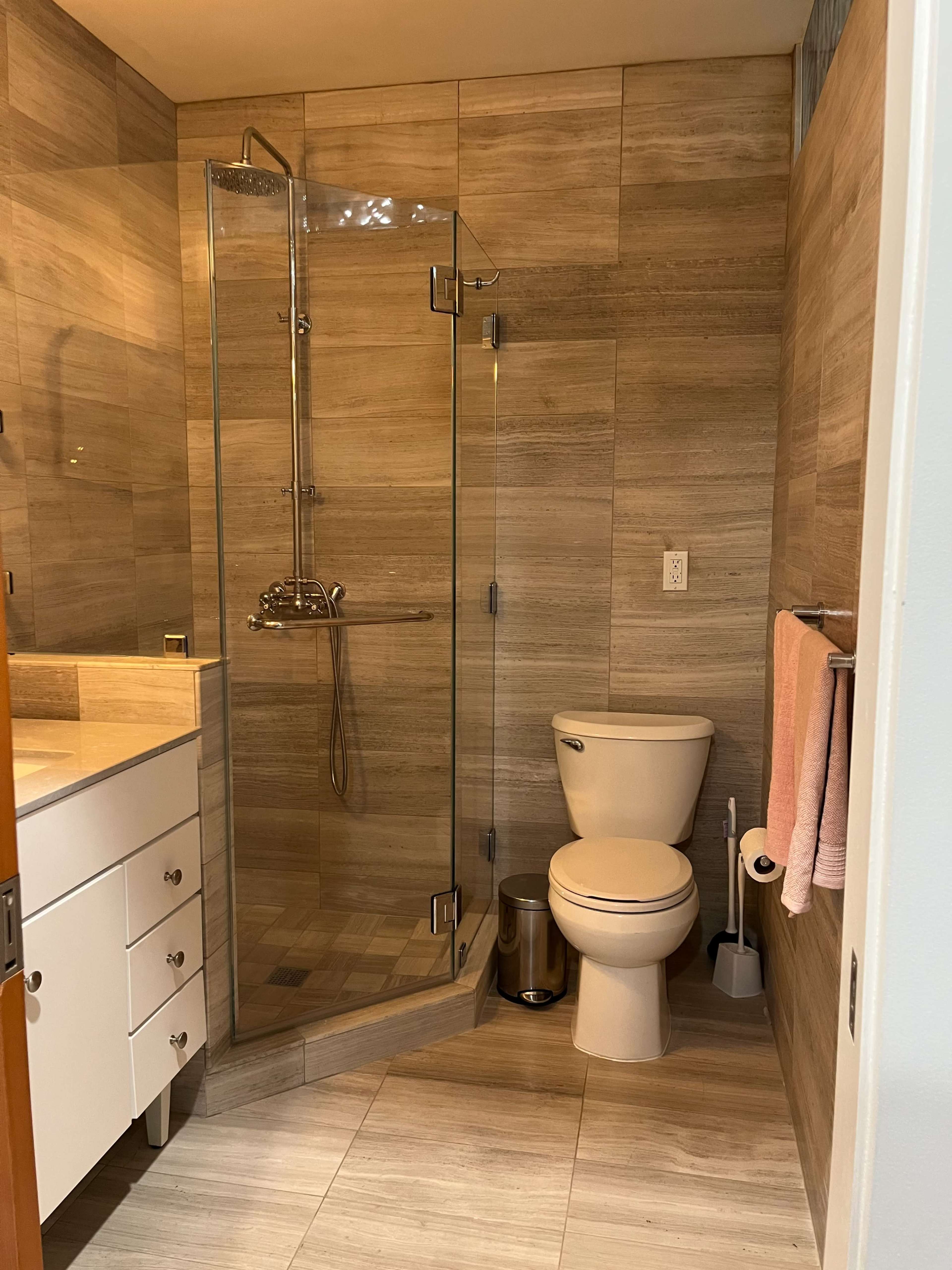 The image shows a modern bathroom featuring a glass shower, a toilet, a sink with drawers, and towel hanging on a rack.