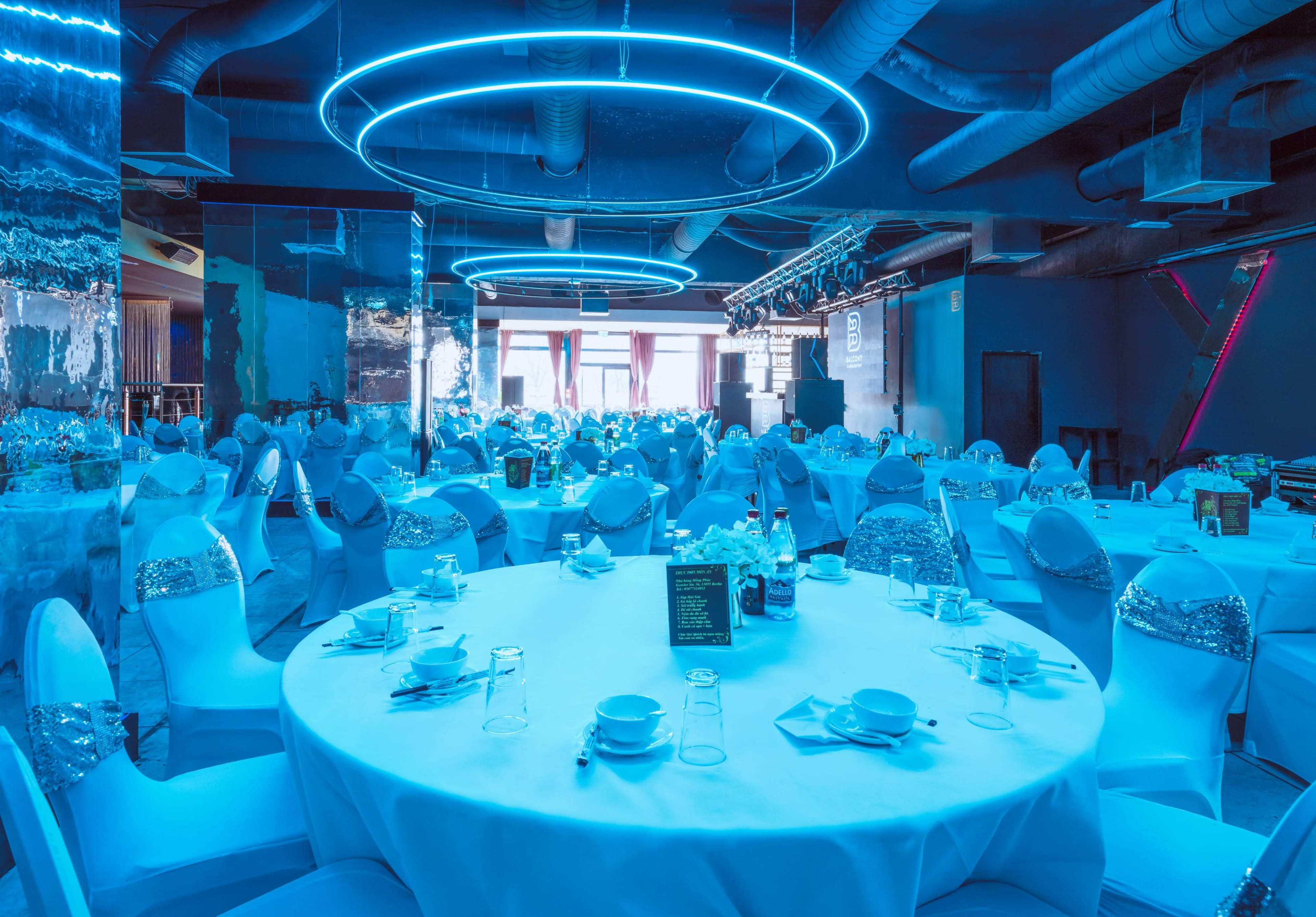 A spacious banquet hall is set up with round tables covered in white tablecloths and decorated with blue lighting.