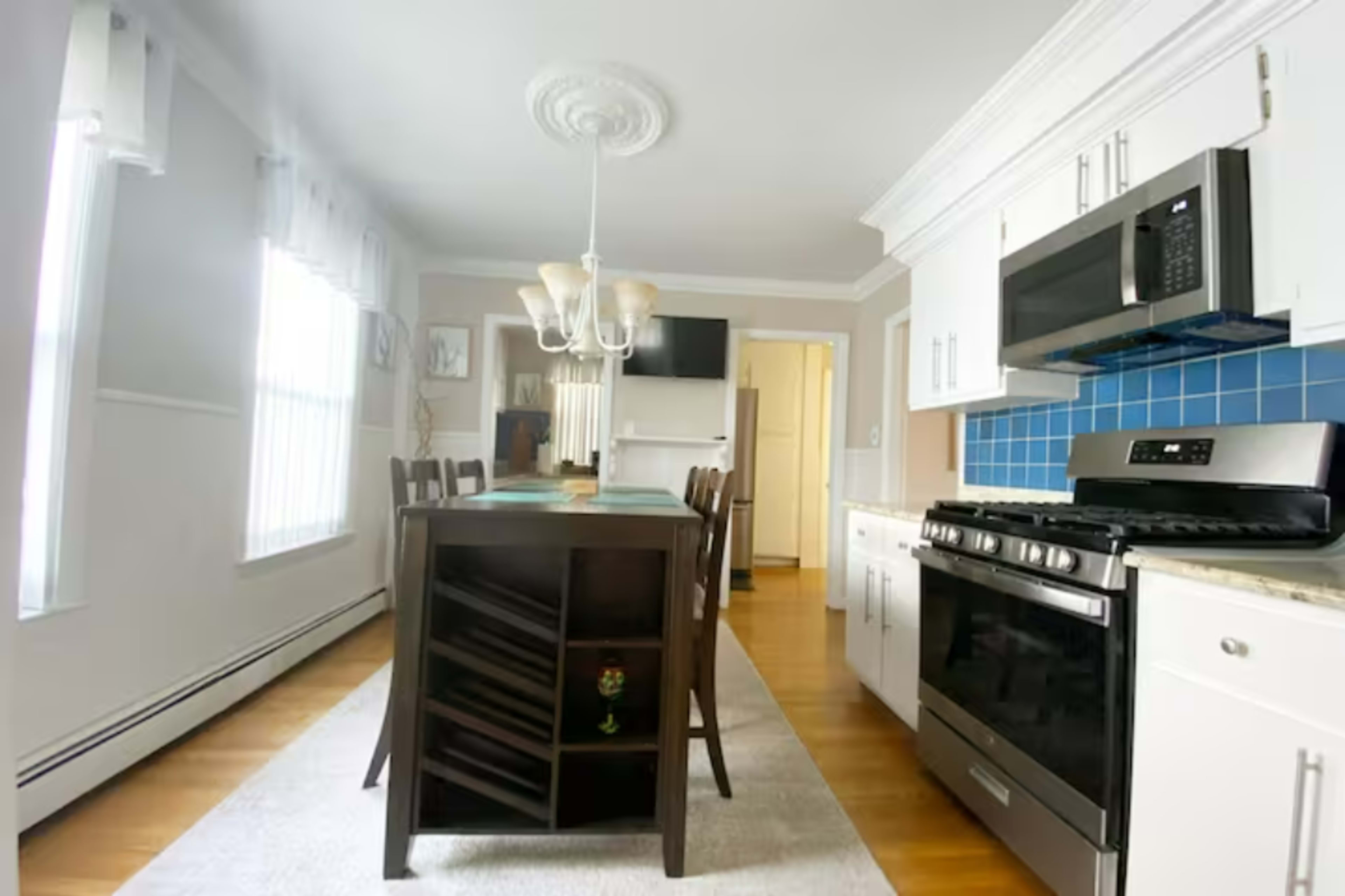 A kitchen with a central dining table, stainless steel appliances, blue tiled backsplash, and natural light coming through window treatments.