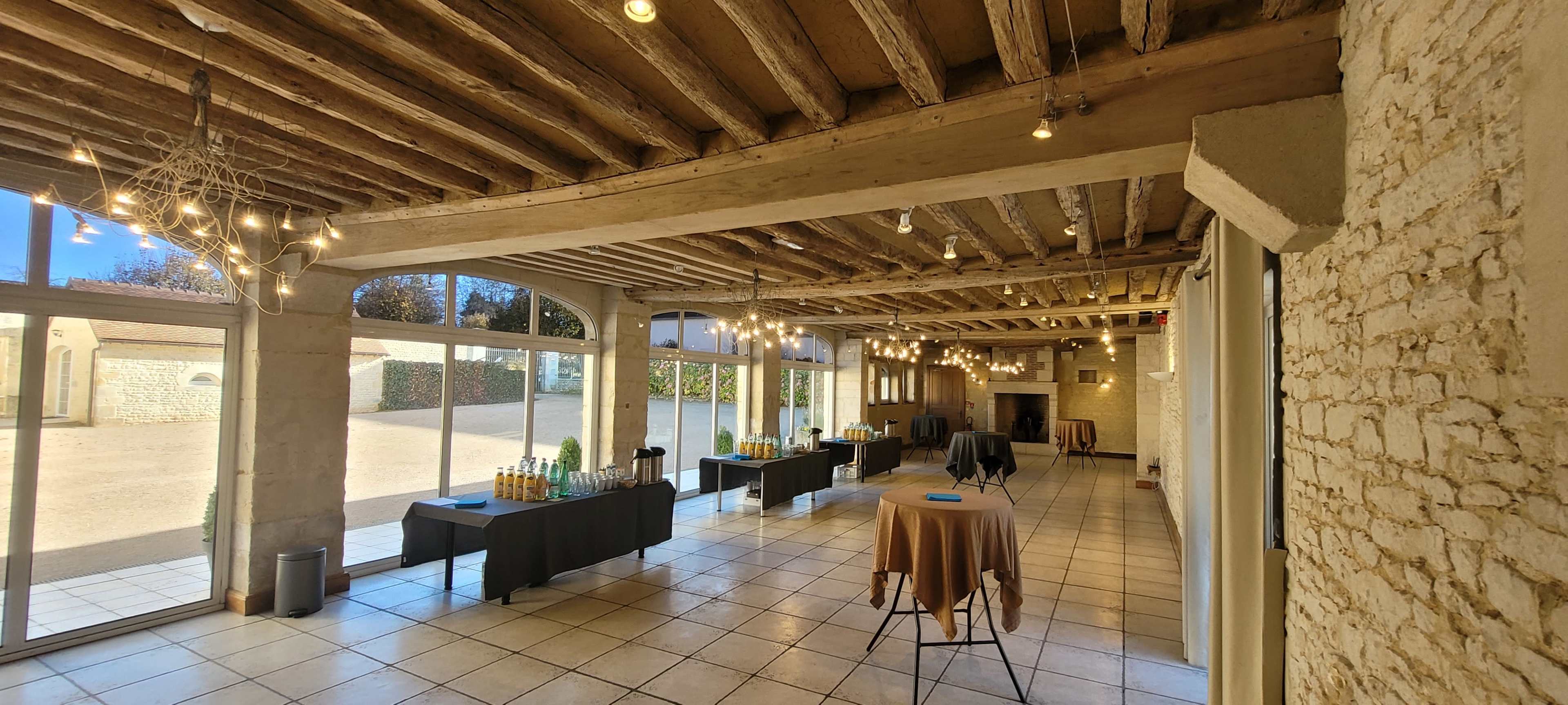 The image shows a spacious event room with wooden beams, featuring several tables with beverages and light fixtures, and large glass doors leading to an outdoor area.