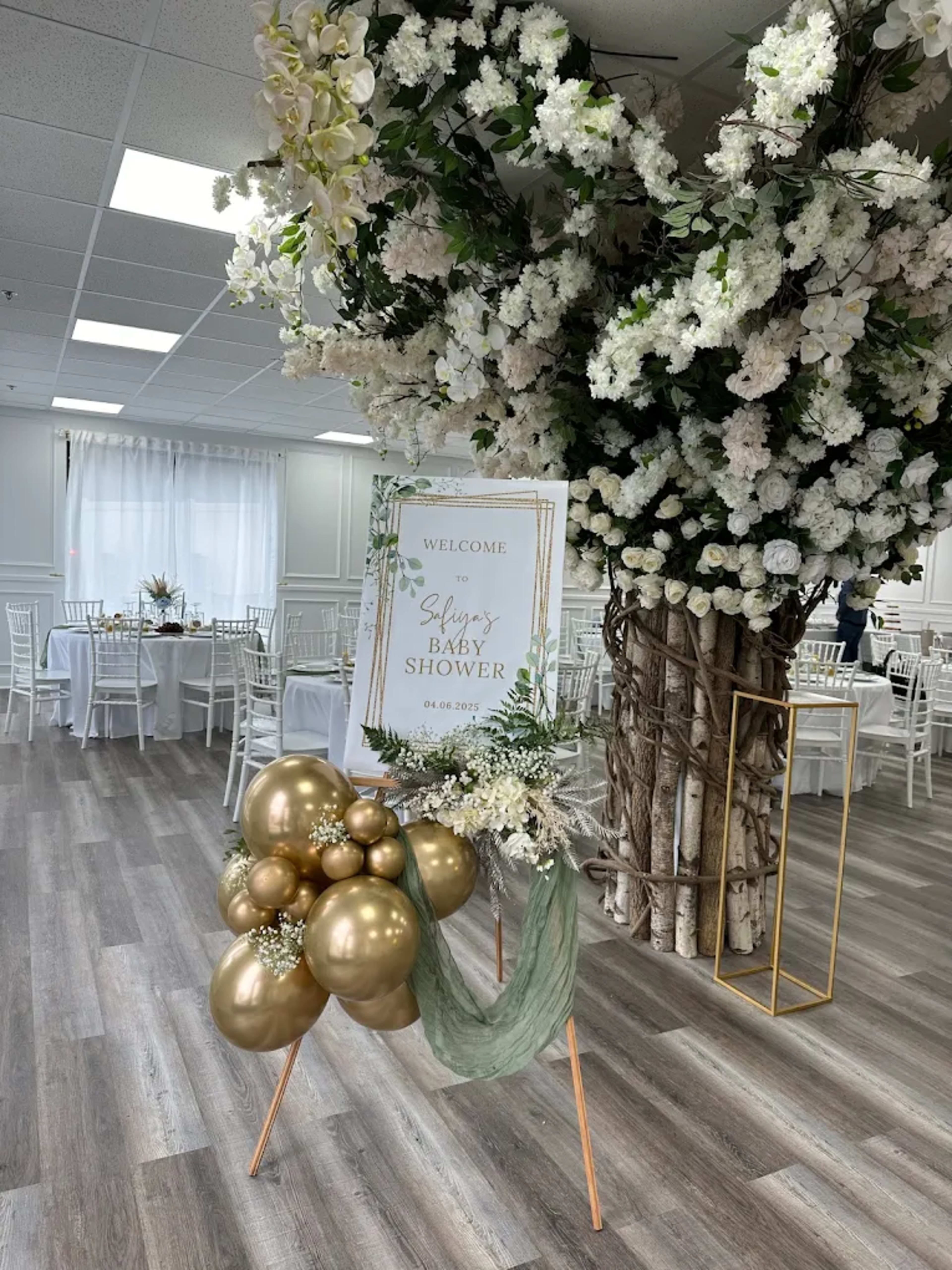 The image shows a decorated event space for a baby shower, featuring a welcome sign amidst floral arrangements and golden balloons.