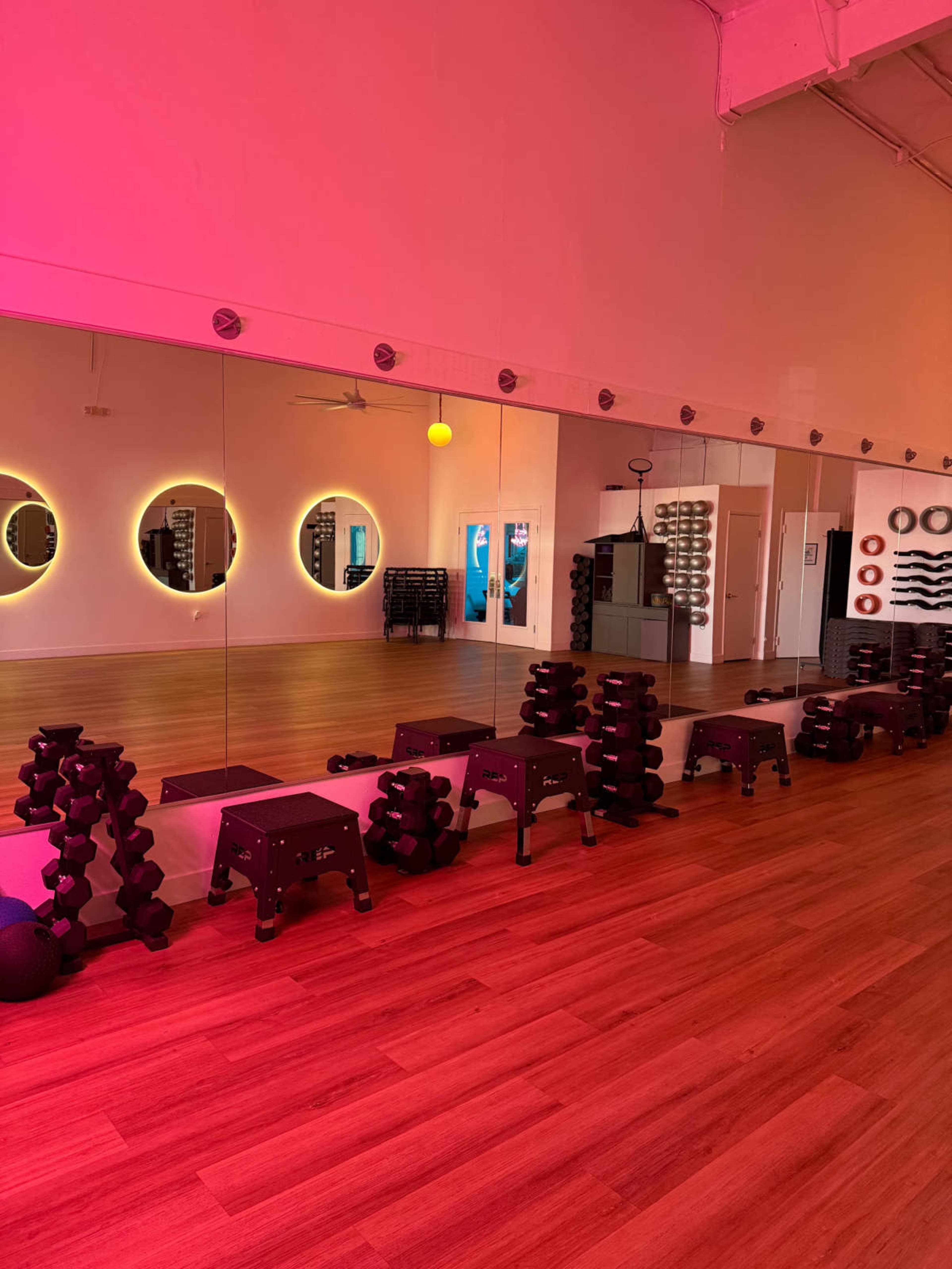 The image shows a modern fitness studio with bright pink lighting, featuring dumbbells and step platforms arranged along a mirrored wall.