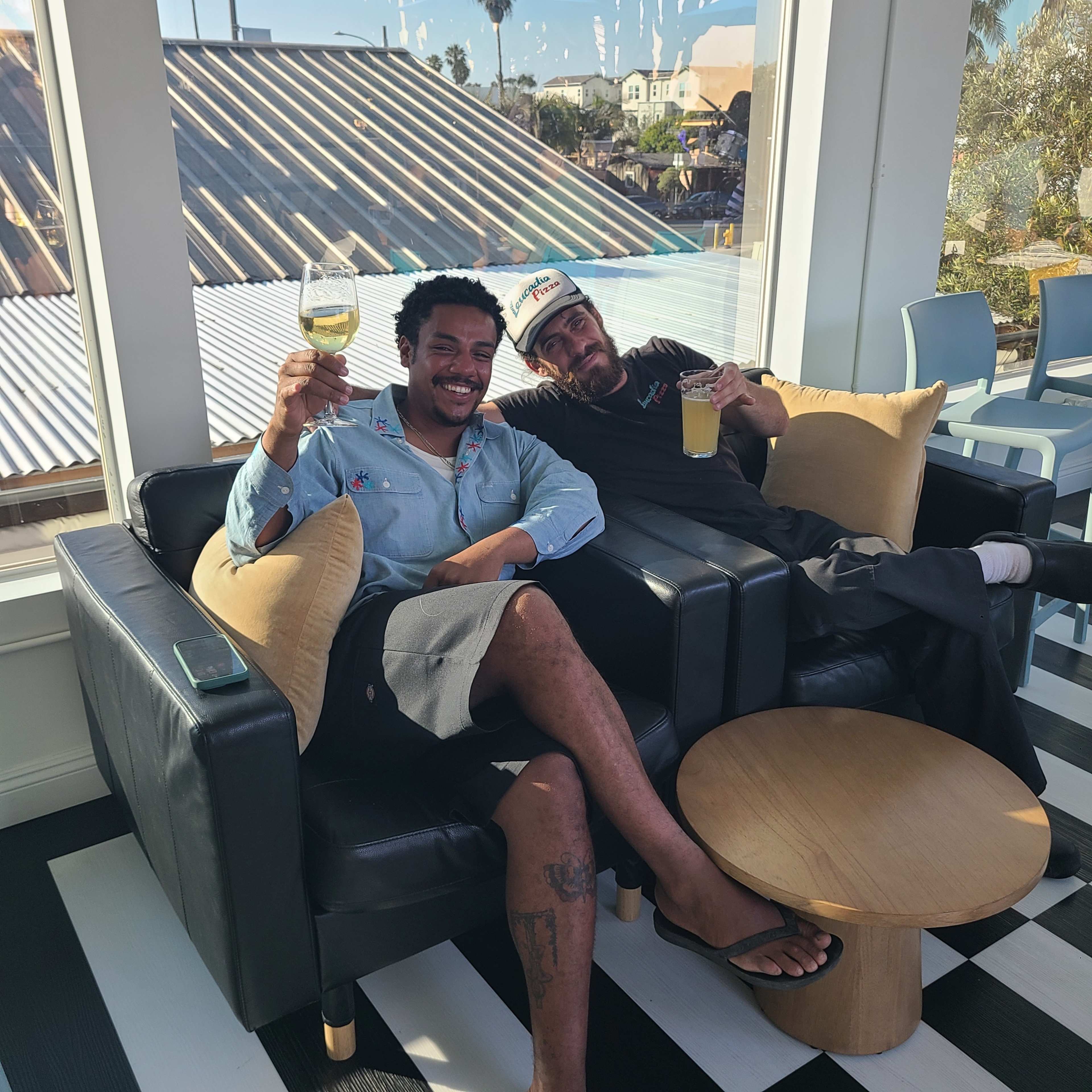 Two men are sitting on a black leather couch in a brightly lit room, both holding drinks while smiling at the camera.