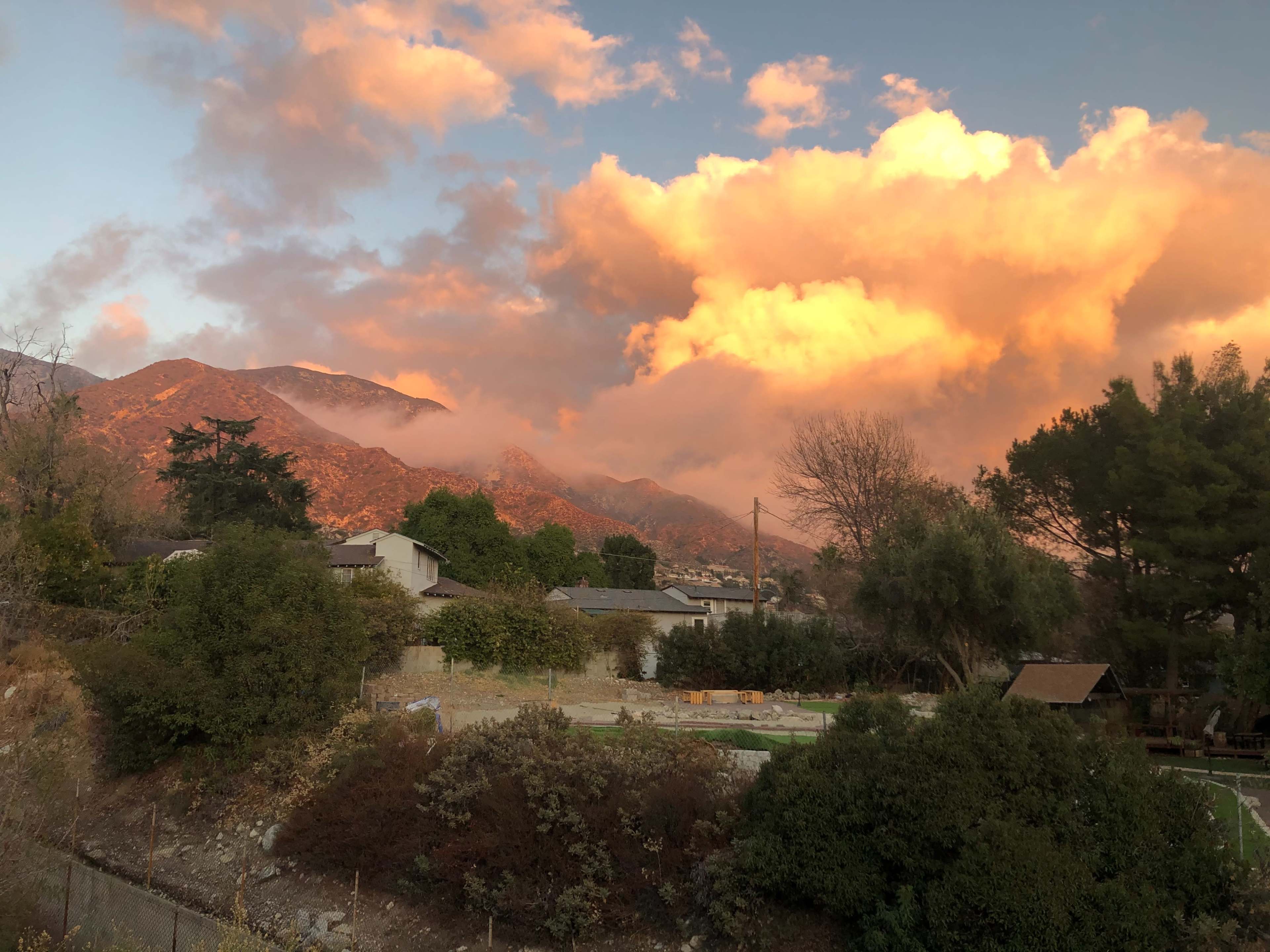 The scene shows a mountain landscape at sunset, with vibrant clouds illuminated by the setting sun above residential buildings and trees.