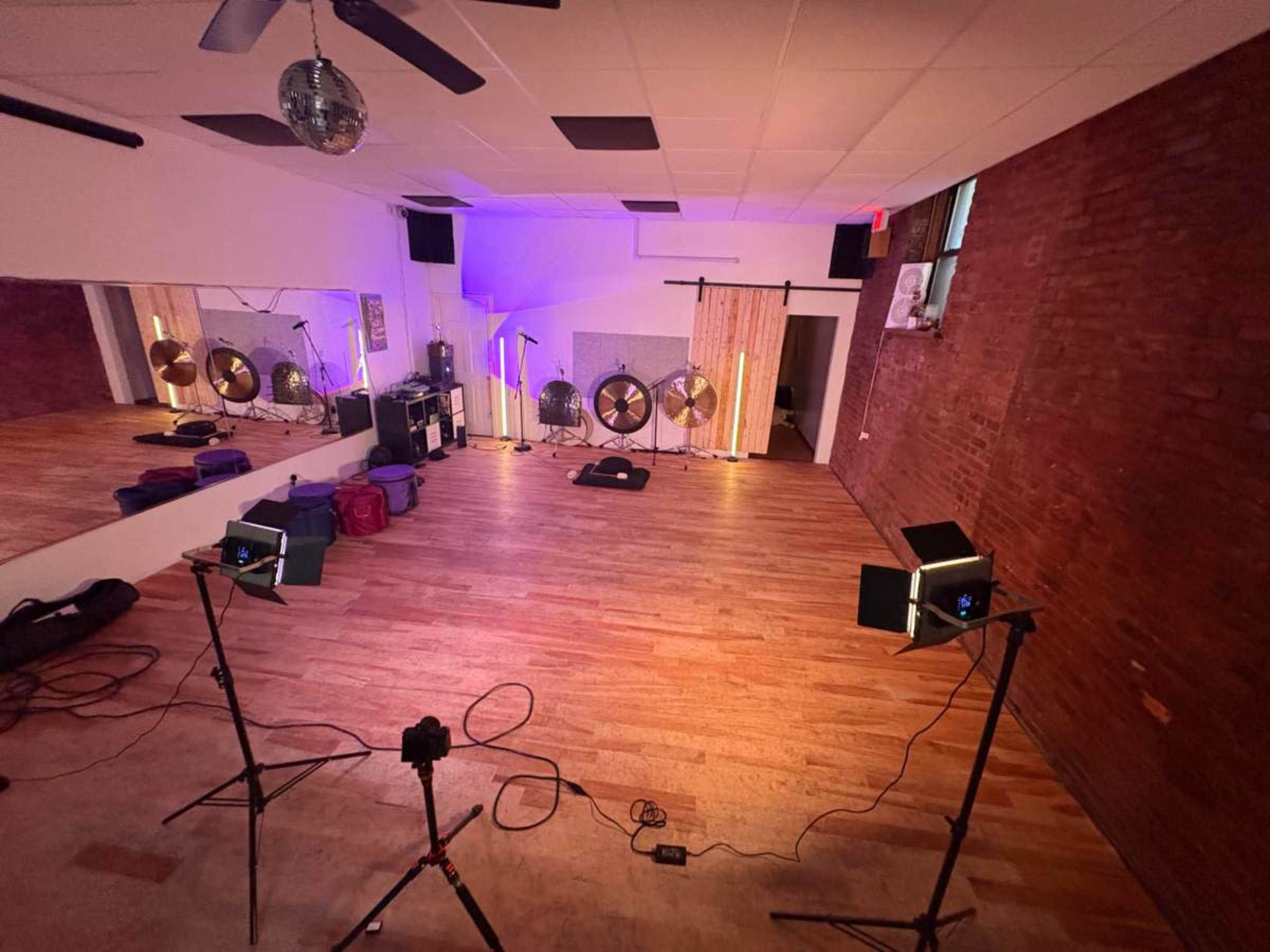A spacious room with wooden flooring features mirrored walls, stage lighting, and musical instruments set up in the background.