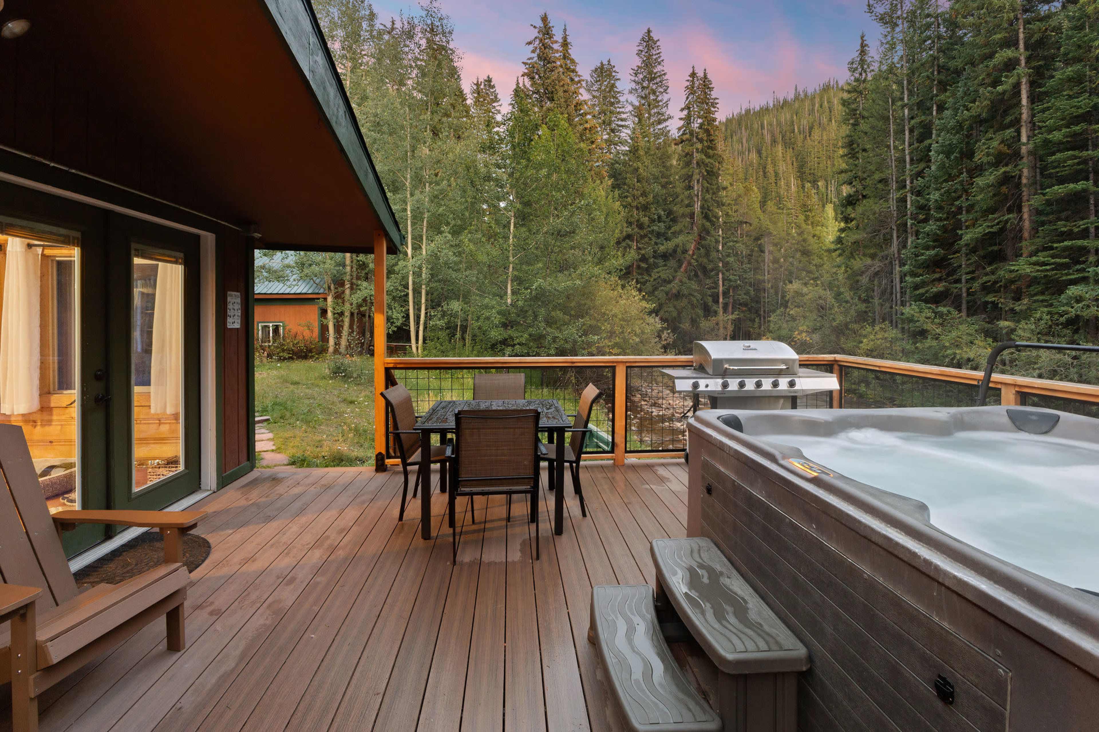 A wooden deck features a hot tub, outdoor dining table, and a barbecue grill, surrounded by tall trees and mountains.
