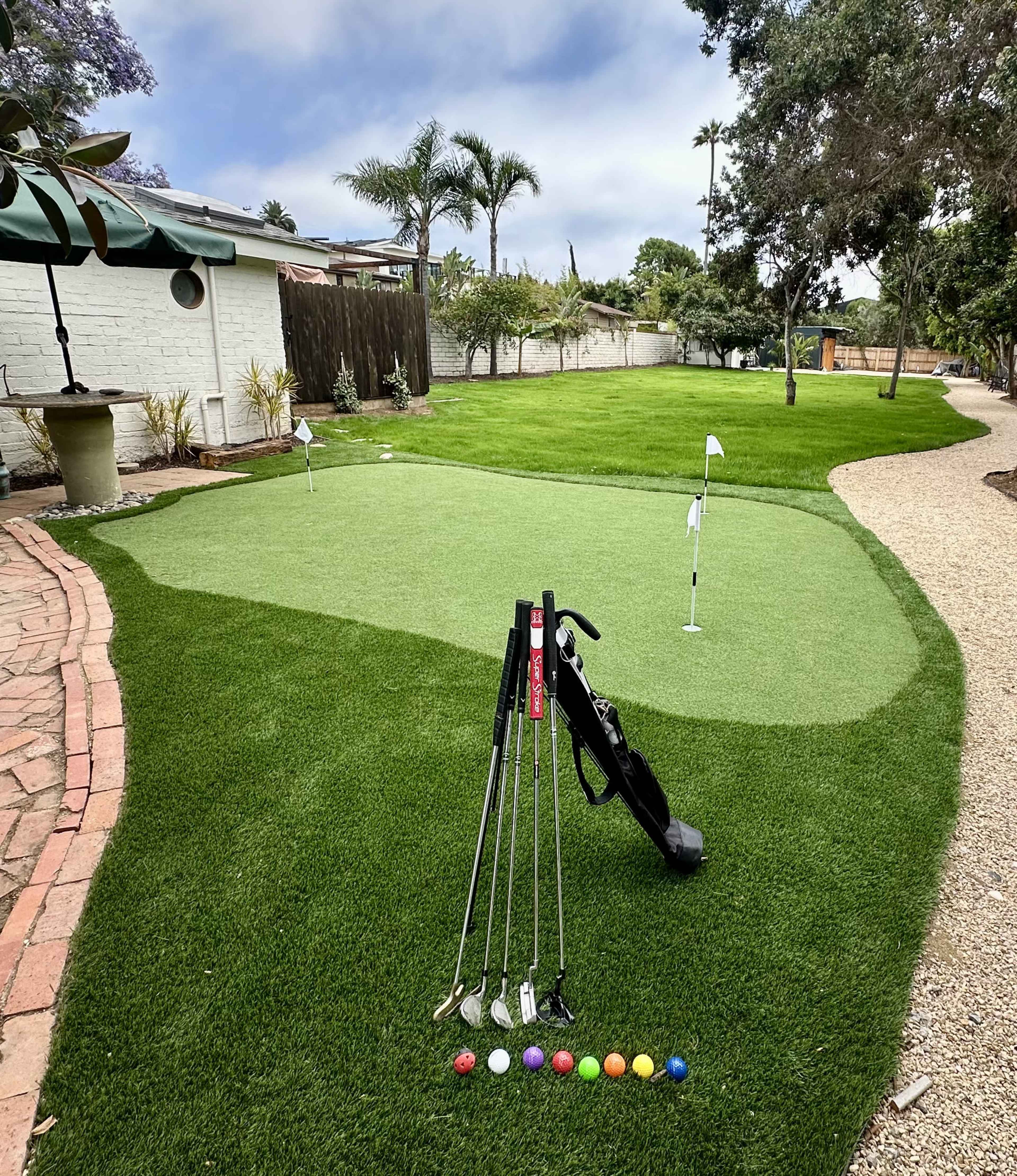 The image shows a putting green with golf clubs and colorful balls arranged in front of it, surrounded by well-maintained grass and landscaping elements.