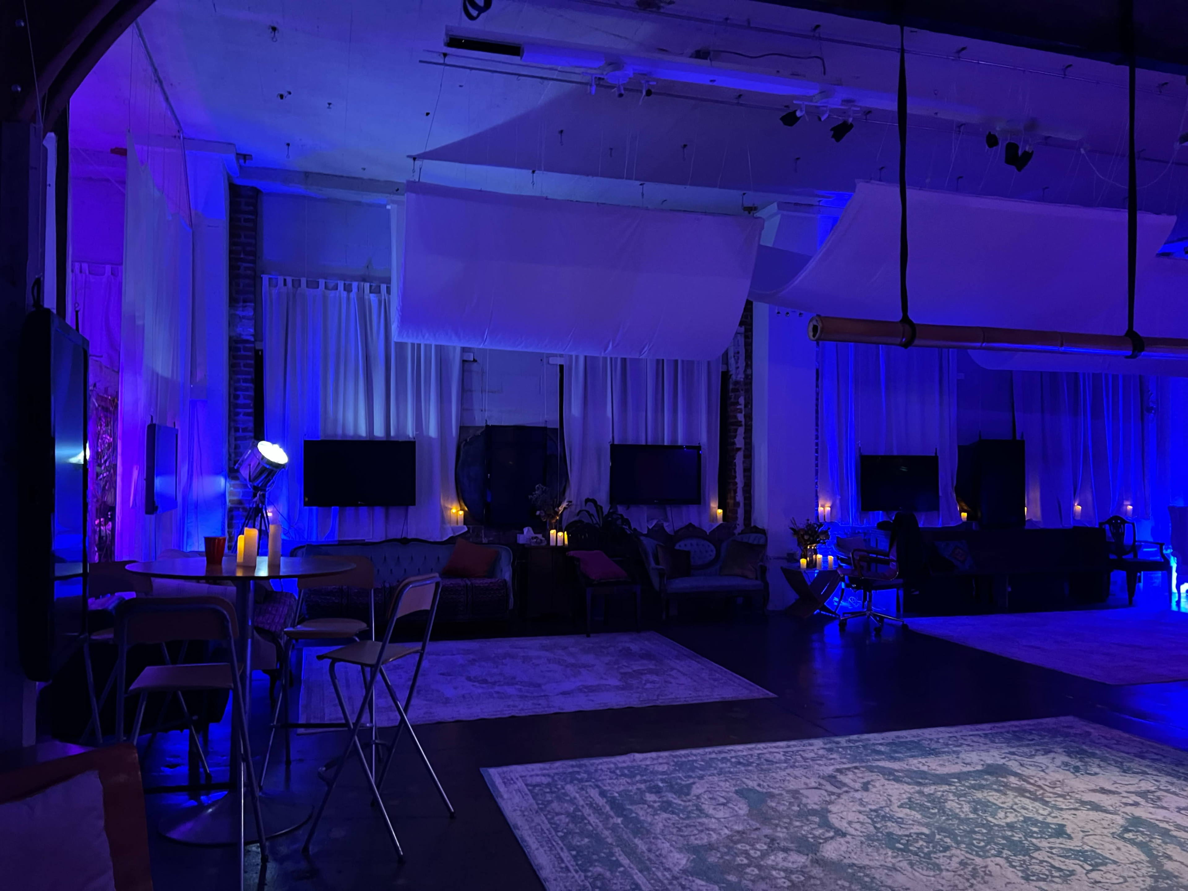The image shows an indoor space with blue lighting, featuring several couches, tables, and screens against a backdrop of white curtains.