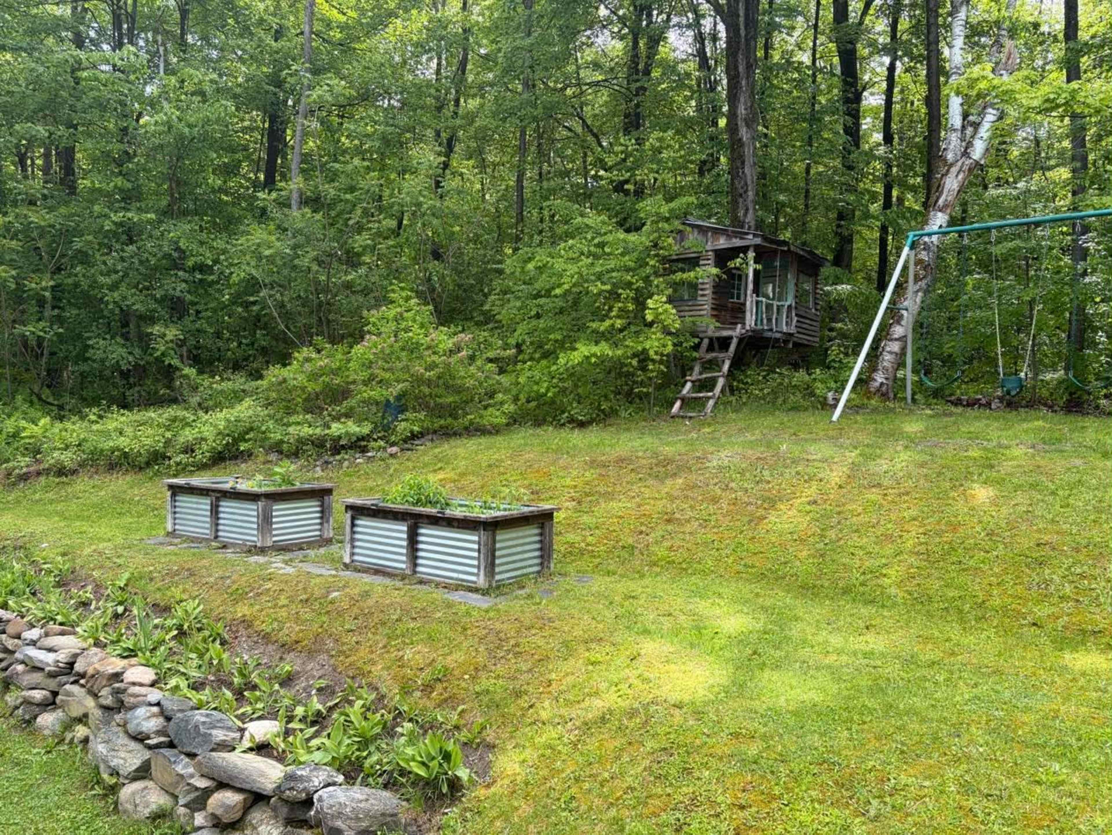 The image shows a lush green yard with two raised garden beds, a wooden playhouse in the background, and a swingset to the right, all surrounded by dense trees.