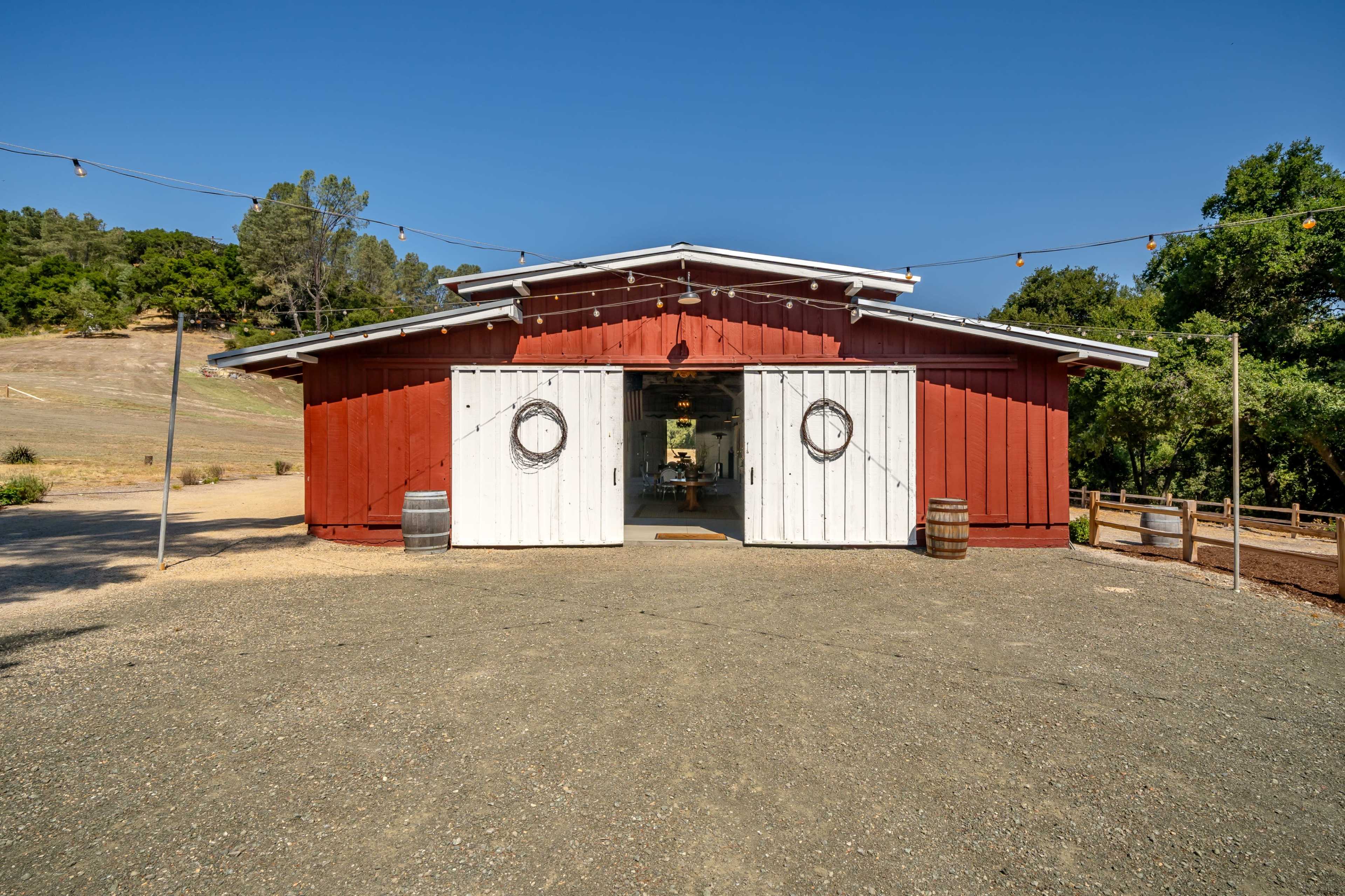 A red barn with white doors stands at the end of a gravel path, surrounded by trees and open landscape under a clear blue sky.
