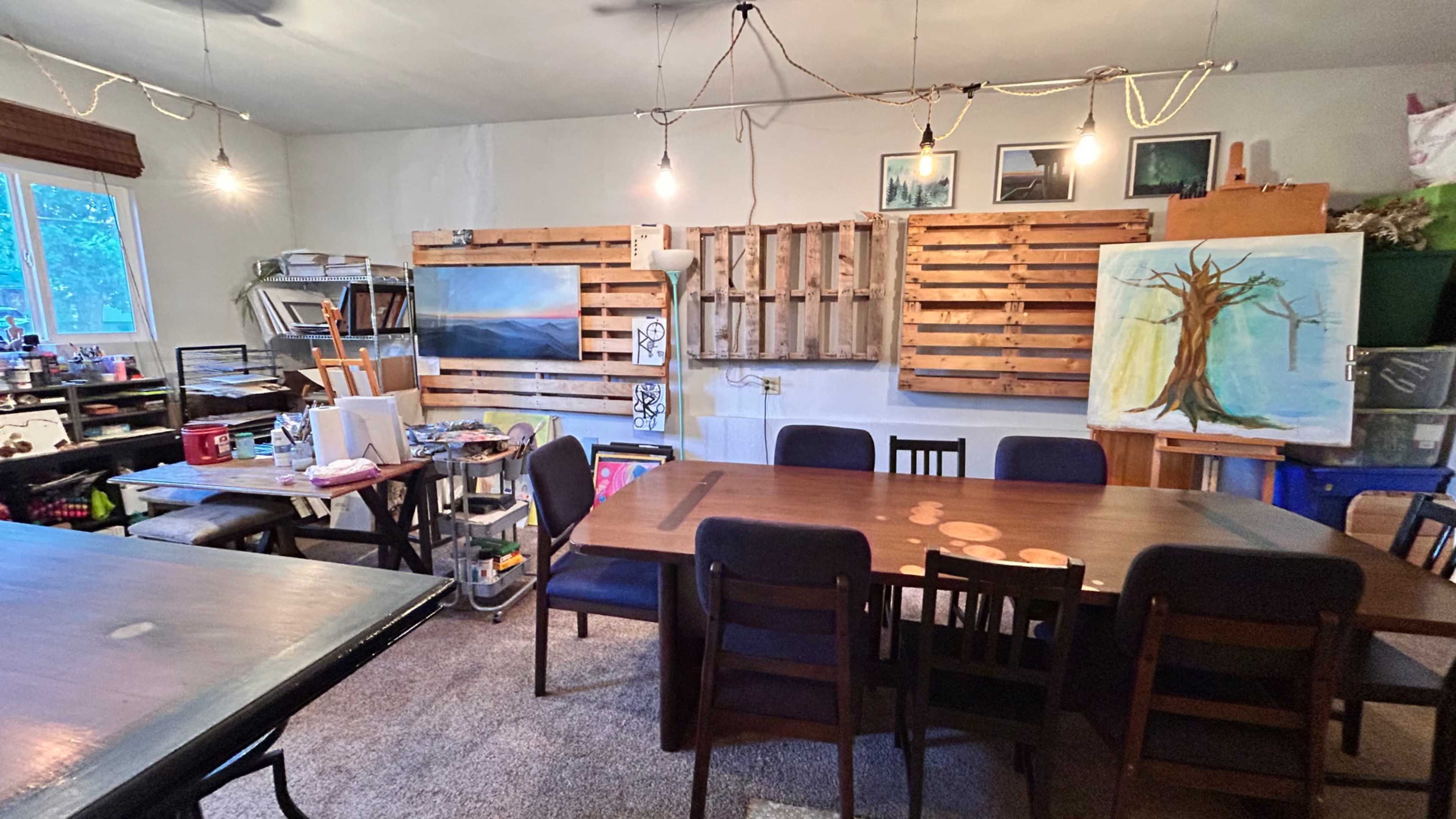 This image shows a creative workspace featuring a large wooden table surrounded by chairs, shelving units with art supplies, and several artworks displayed on the walls.