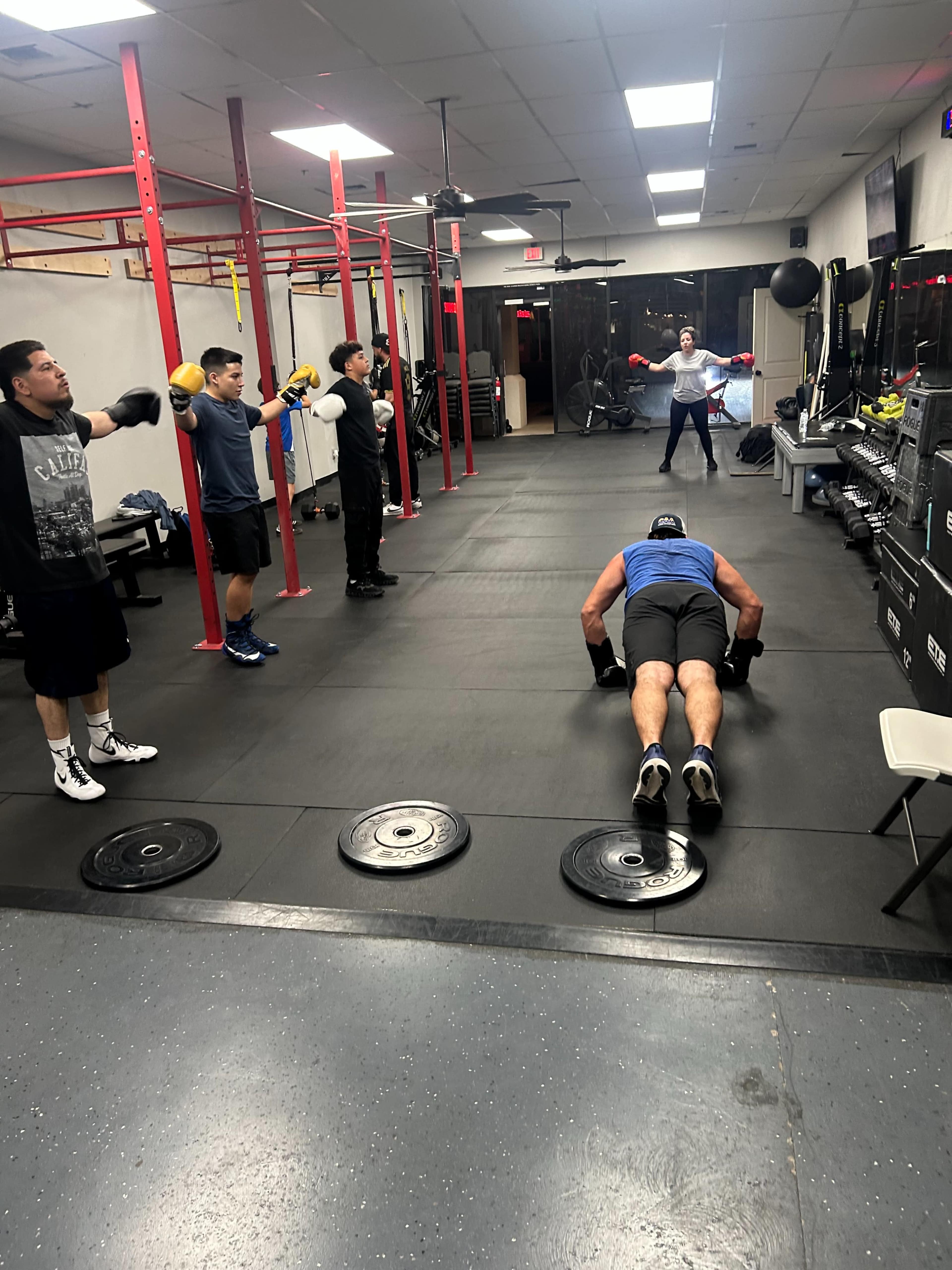 A group of six individuals is engaged in a workout session in a gym, with some practicing boxing techniques and one person doing push-ups on the floor.