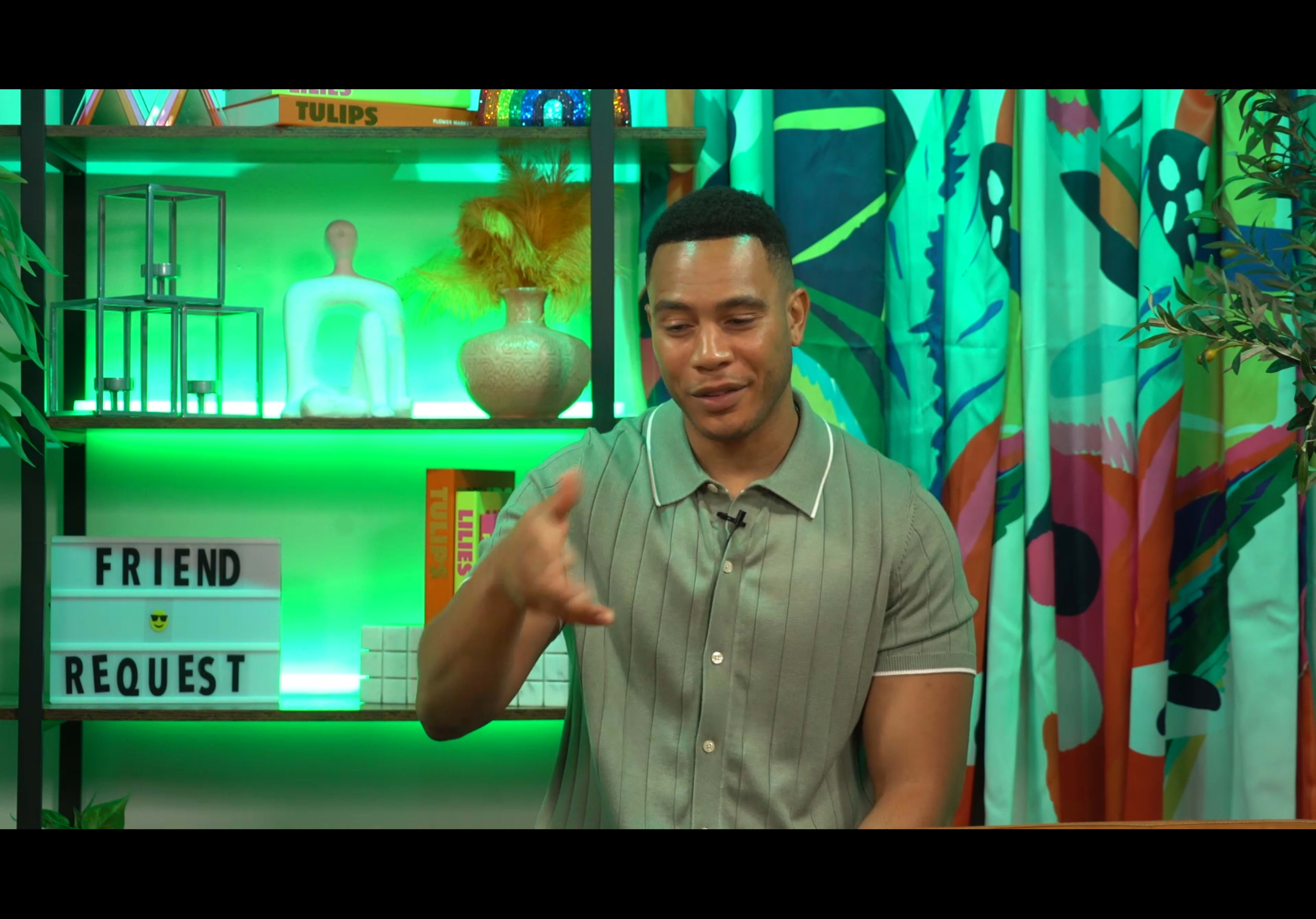 A man in a short-sleeve, collared shirt gestures while sitting in front of a brightly colored backdrop with decorative shelves.
