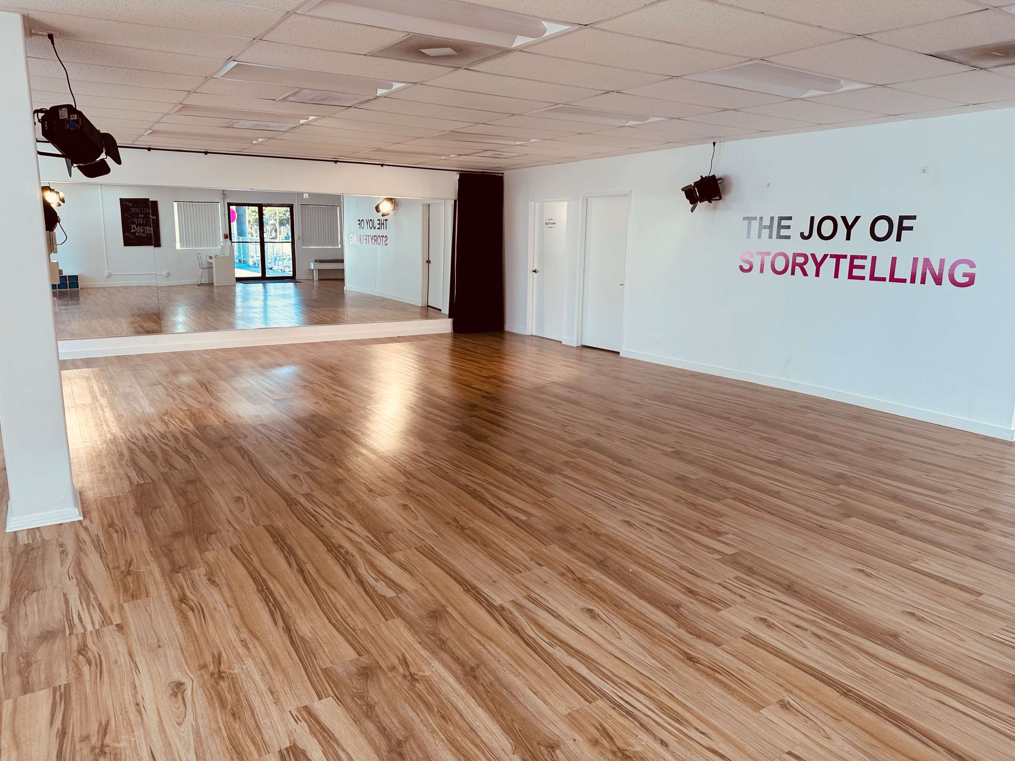 A spacious, empty room with wooden flooring, featuring a large mirror on one wall and the phrase "THE JOY OF STORYTELLING" displayed on another.