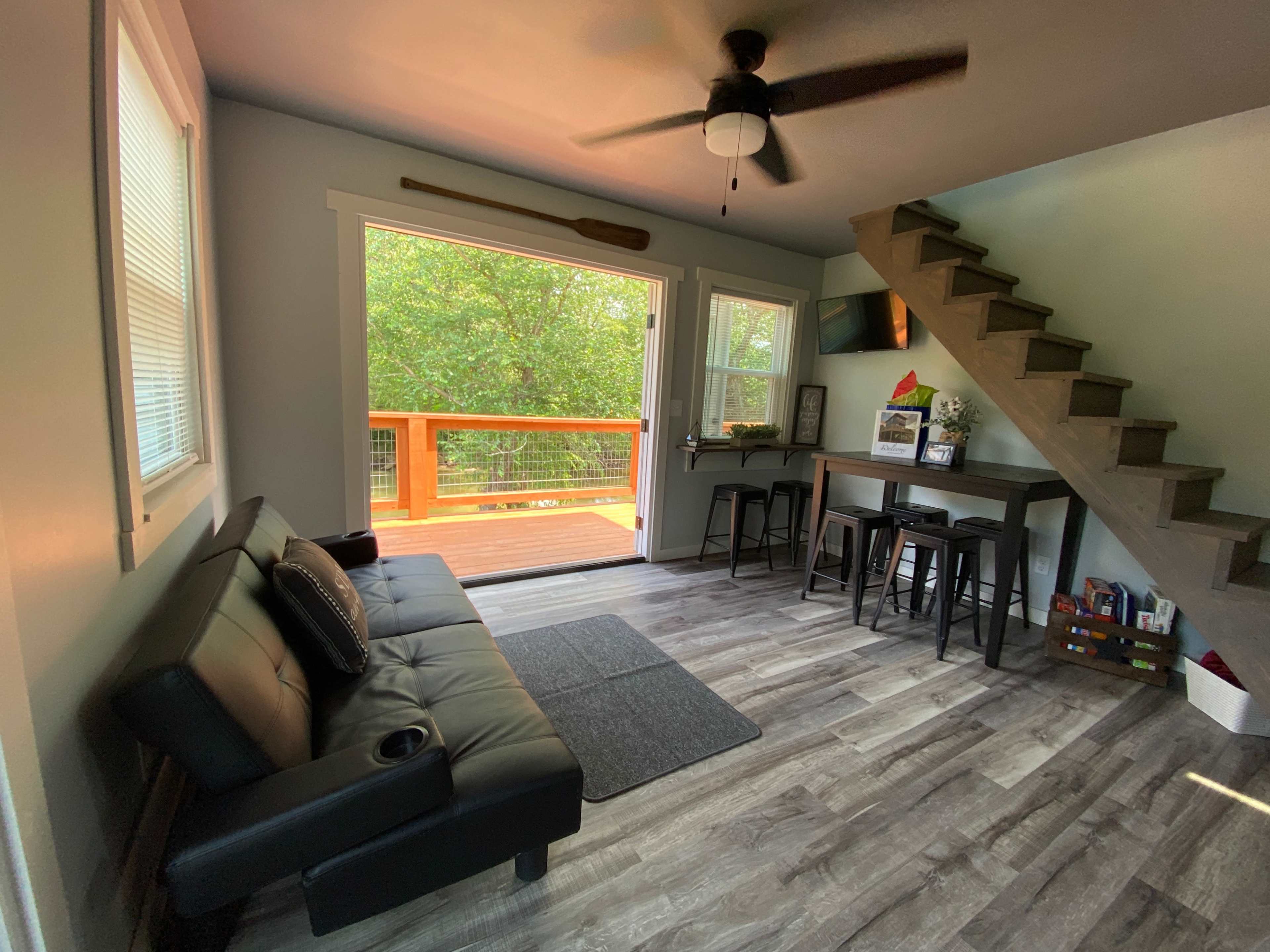 The image shows a modern living area with a black futon, a small dining table, and a staircase leading to an upper level, with large windows overlooking a deck.