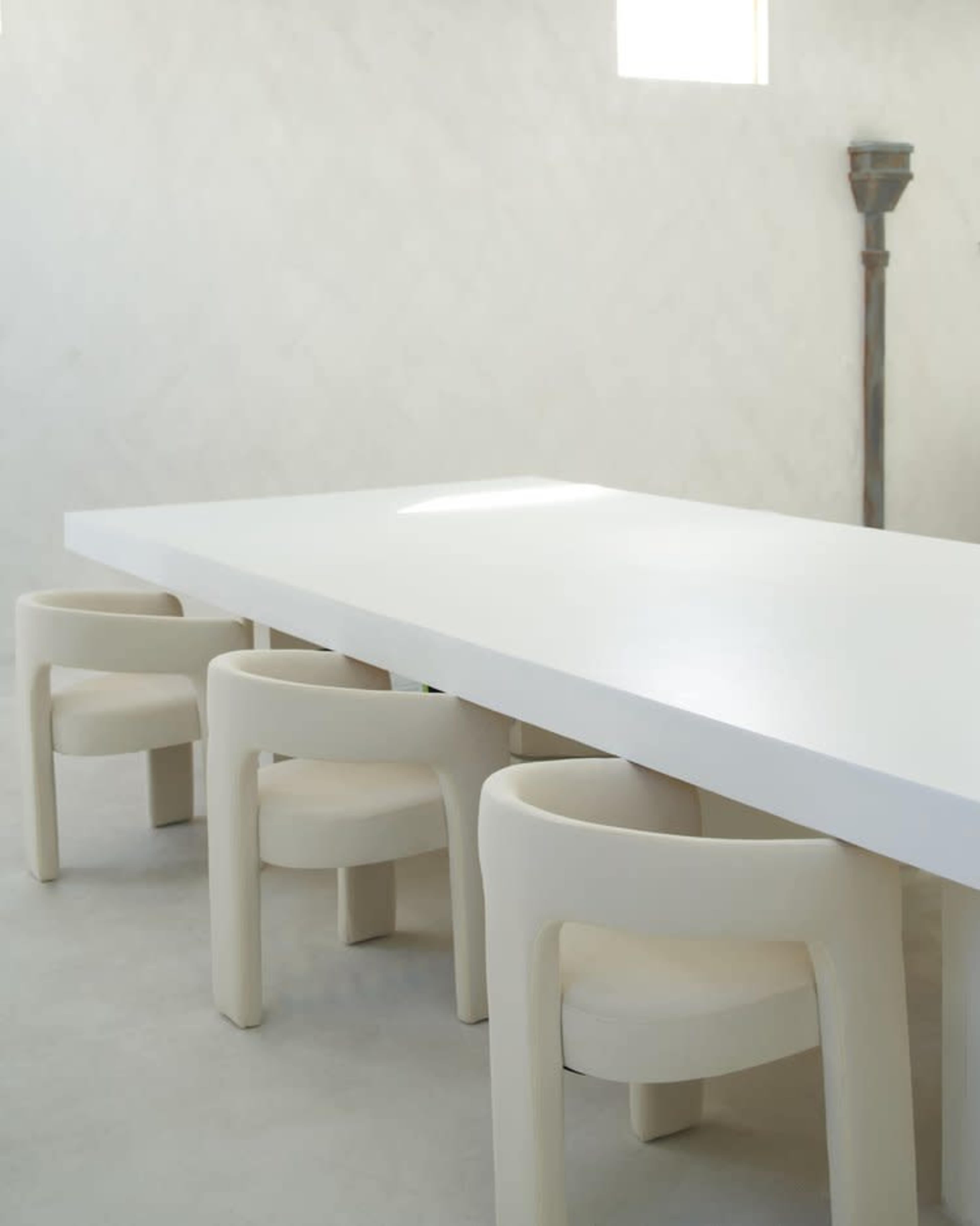 A long white table is surrounded by four white, circular chairs in a minimalist room with light-colored walls.