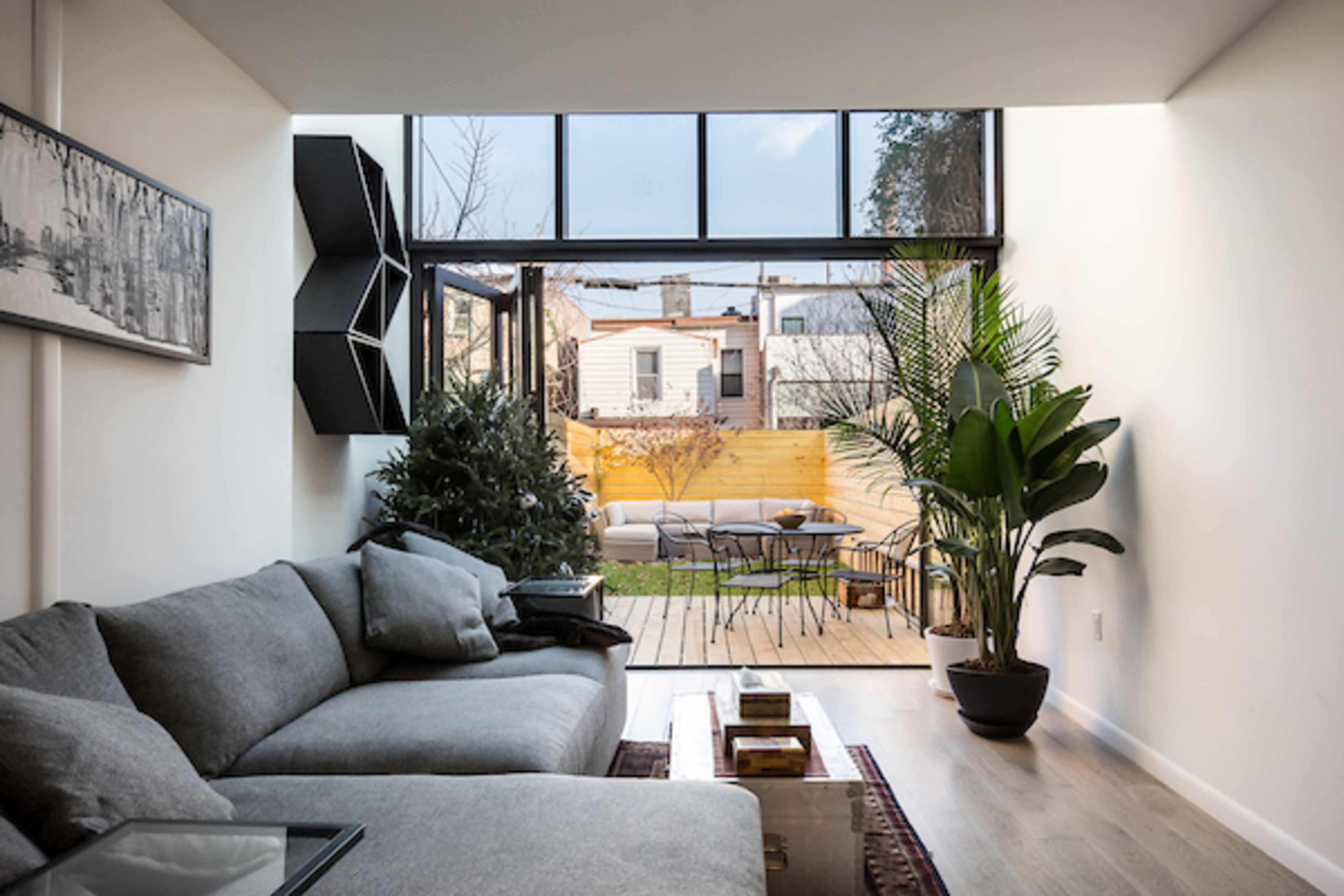 A modern living room features a gray sofa, large windows that open to a backyard, and a view of patio seating and greenery.