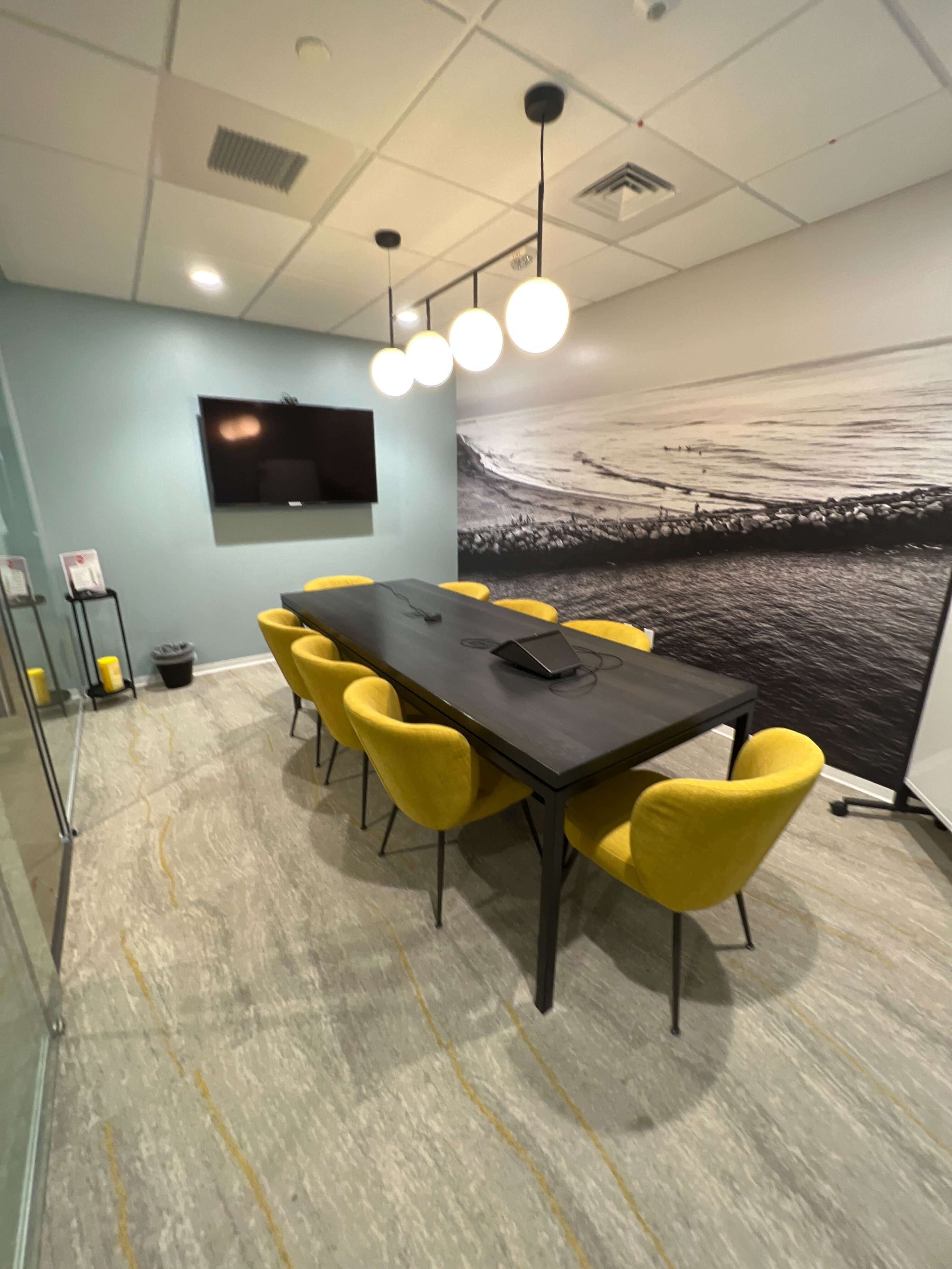 A conference room features a large dark table surrounded by yellow chairs, with a television mounted on the wall and soft lighting from pendant fixtures.