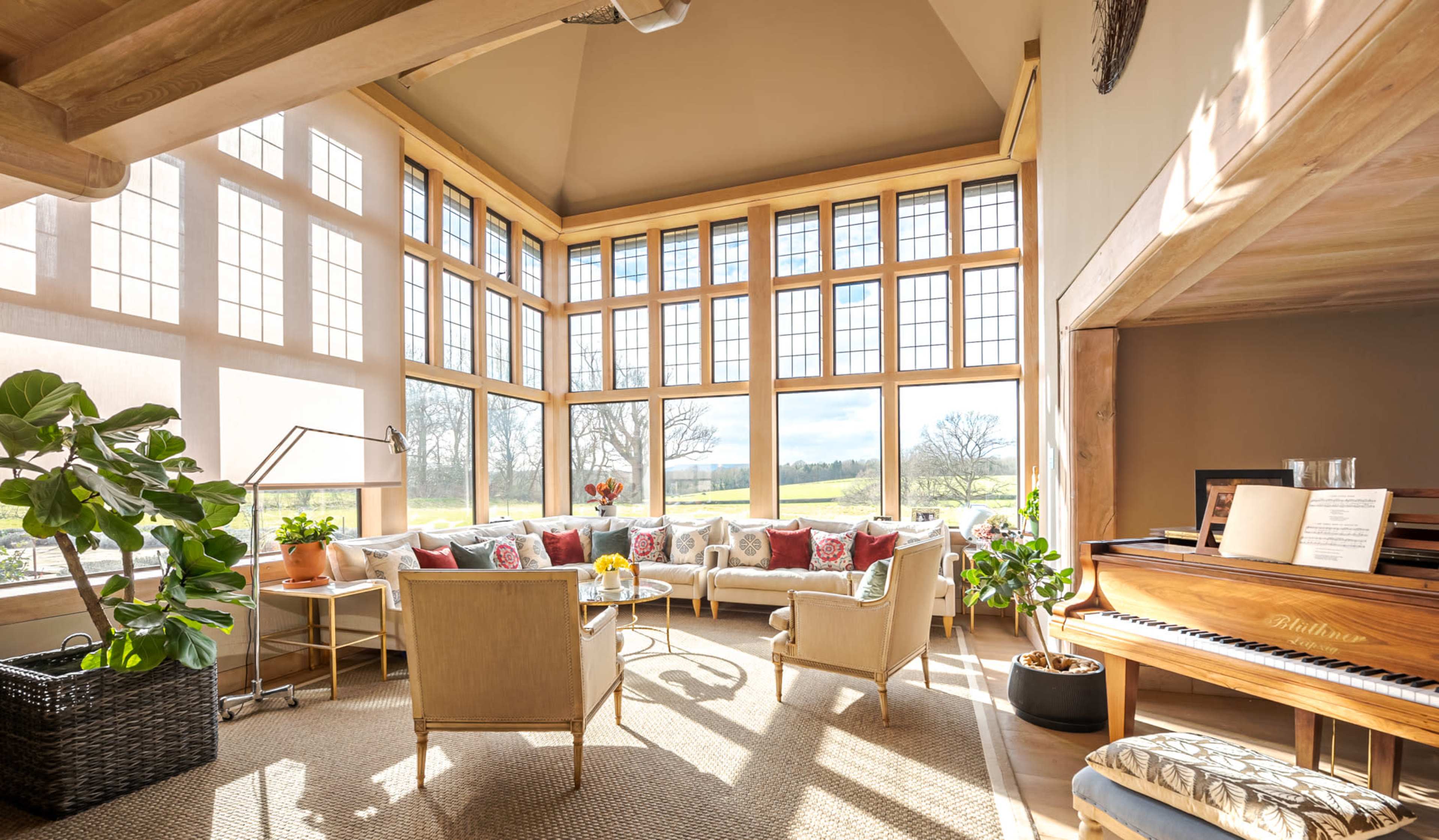 A sunlit living room features large windows, a curved sofa with colorful cushions, two chairs, a piano, and indoor plants.