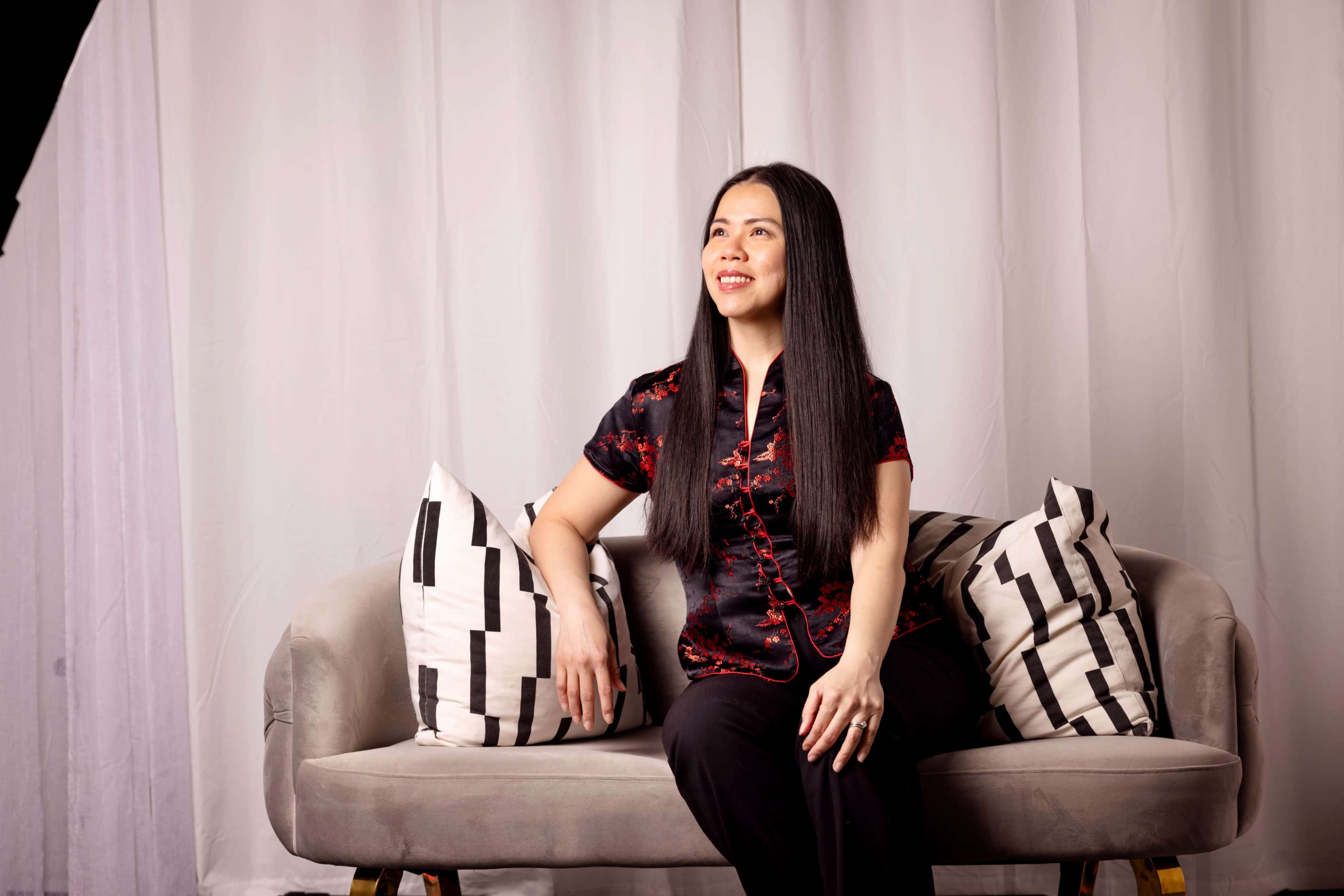 A woman sits on a sofa with patterned cushions, facing slightly to the side against a backdrop of white drapes.