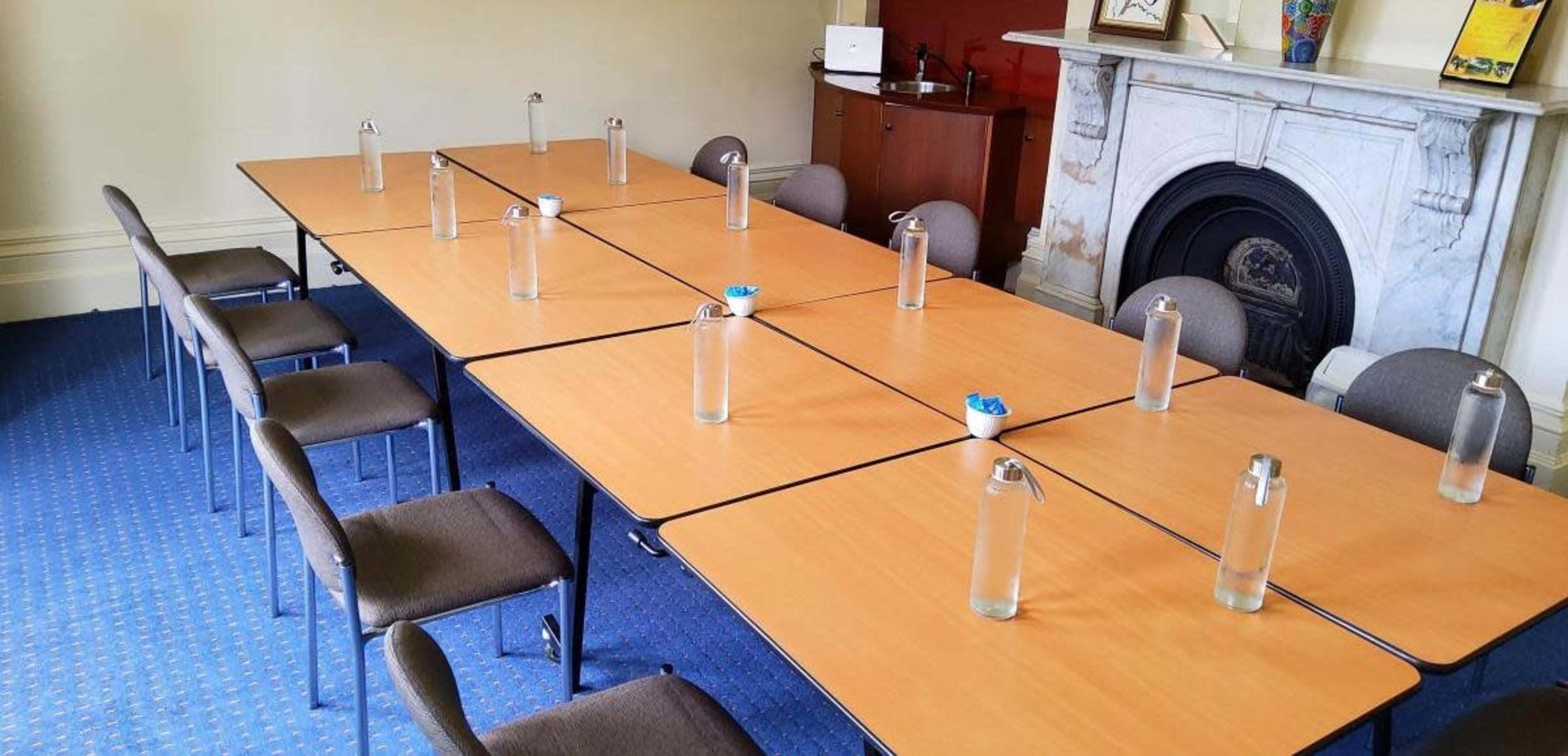 A meeting room is set up with several tables arranged in a U-shape, each containing a glass water bottle and chairs positioned around them.