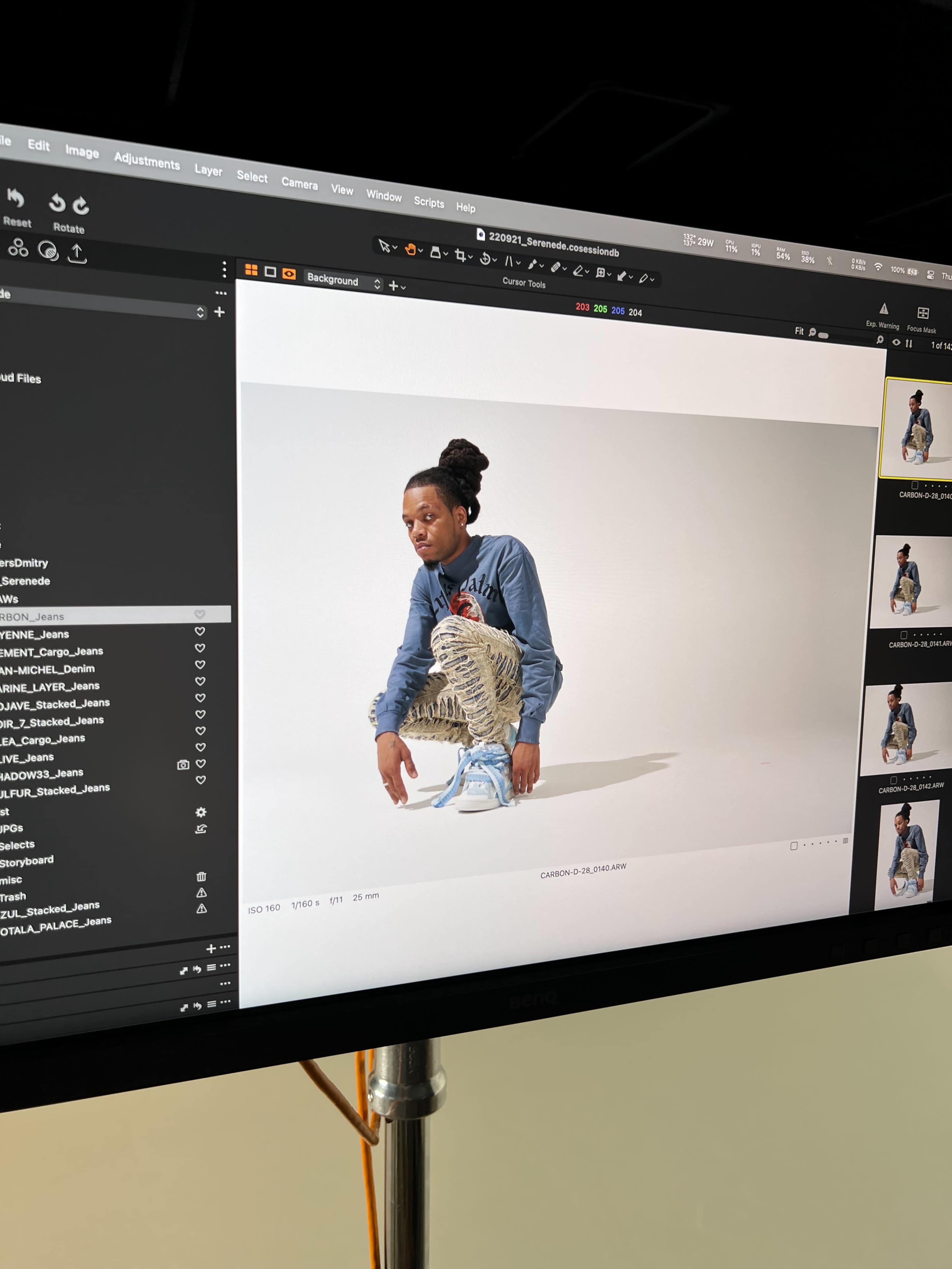 A person wearing a denim jacket and patterned pants is crouching on a white backdrop, with a photography editing interface visible on the screen beside them.