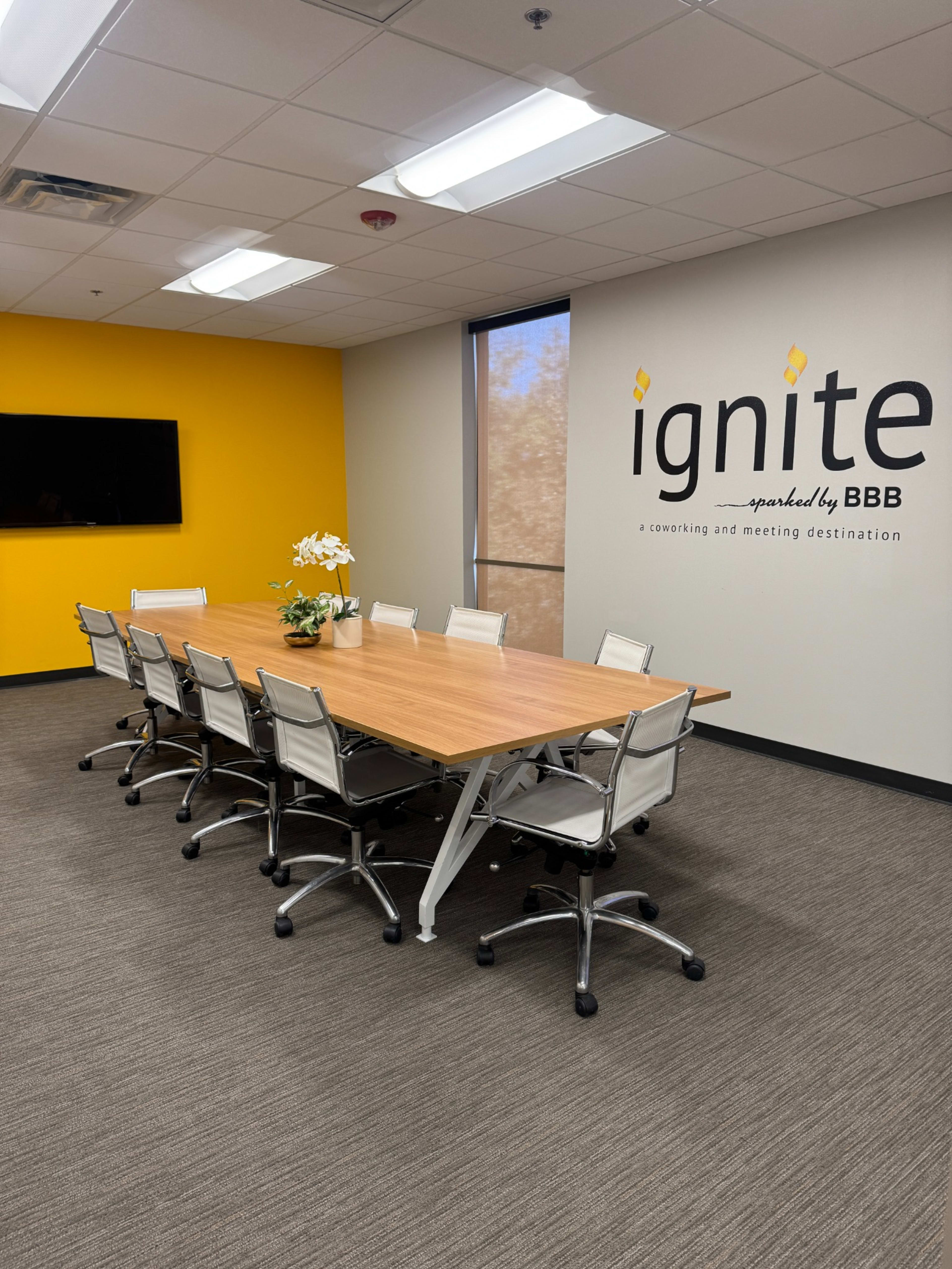A conference room features a large wooden table surrounded by eight swivel chairs, with a bright yellow wall displaying the word "ignite" and a television mounted on the opposite wall.