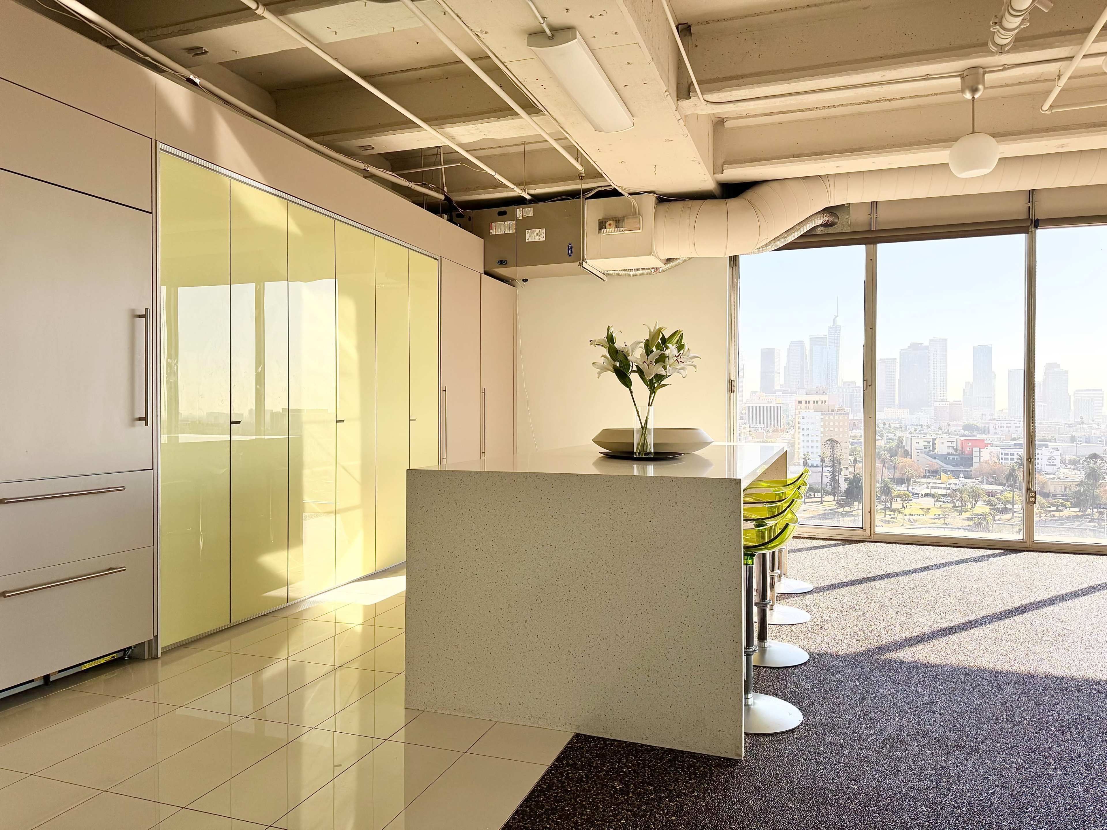 A modern office space features a sleek kitchen island with bar stools and large windows overlooking a city skyline.