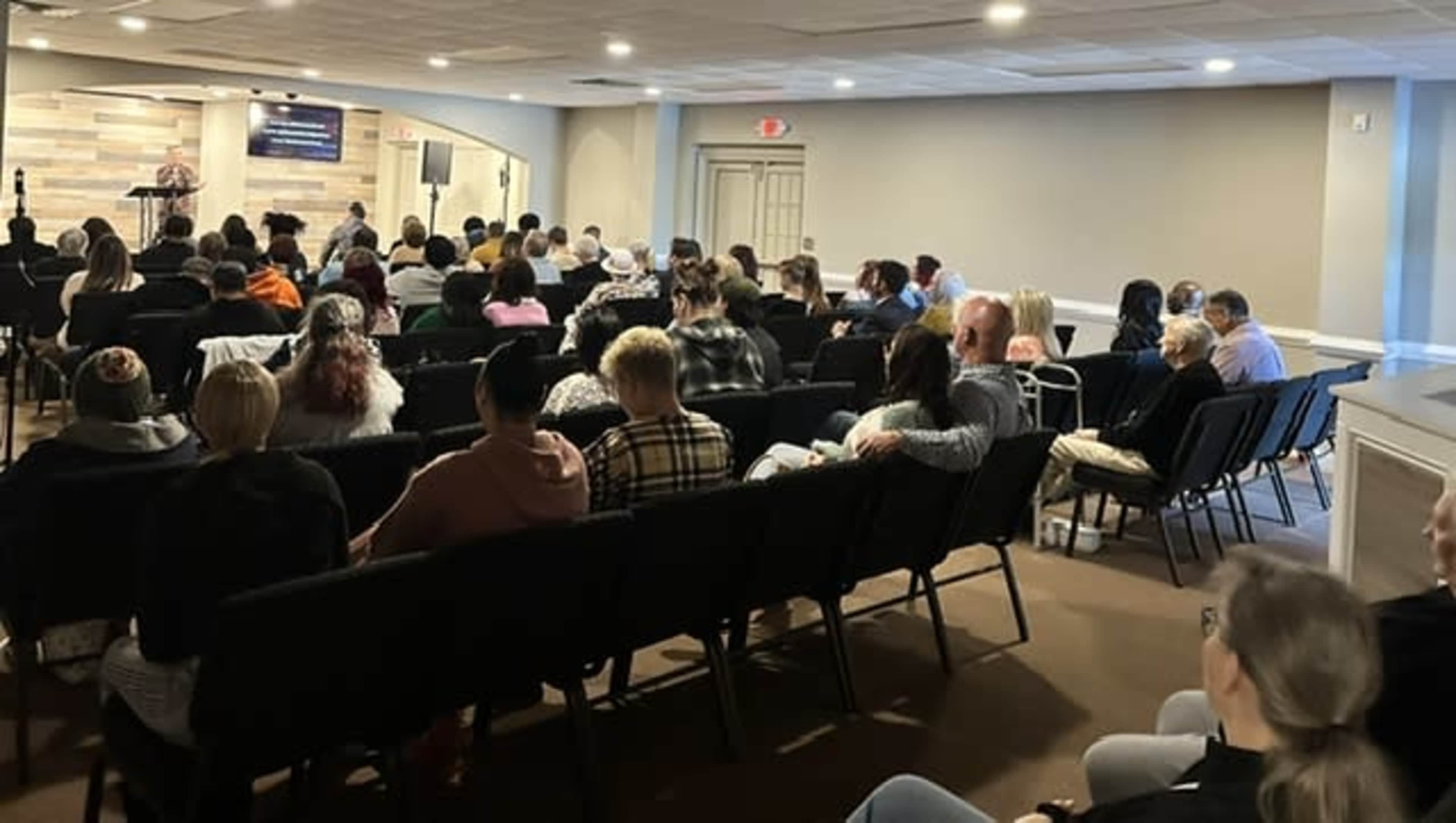 A large audience sits in rows of chairs facing a speaker at a front podium in a well-lit meeting room.