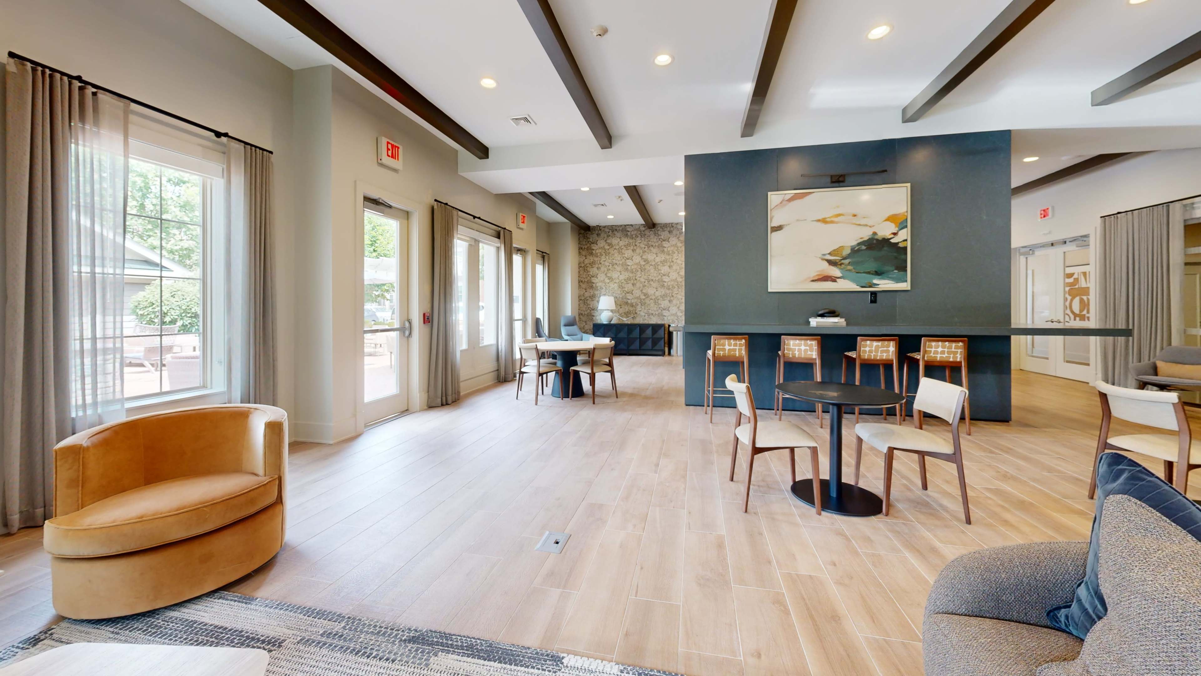 The image shows a spacious room with wooden floors, modern furniture, and large windows that allow natural light to fill the area.