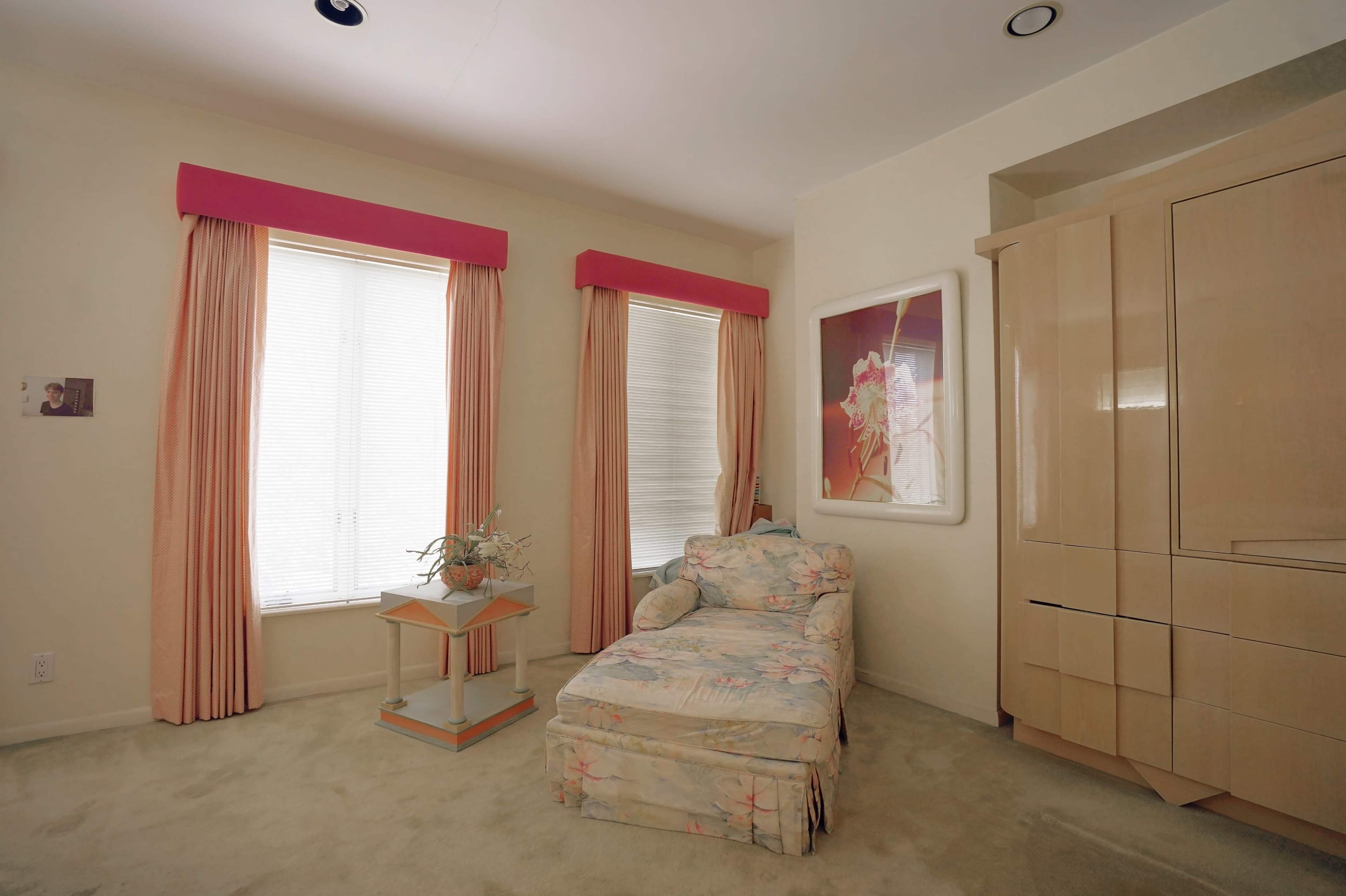 The room features a chaise lounge with floral upholstery, surrounded by two tall windows with pink valances and a tall cabinet against the wall.