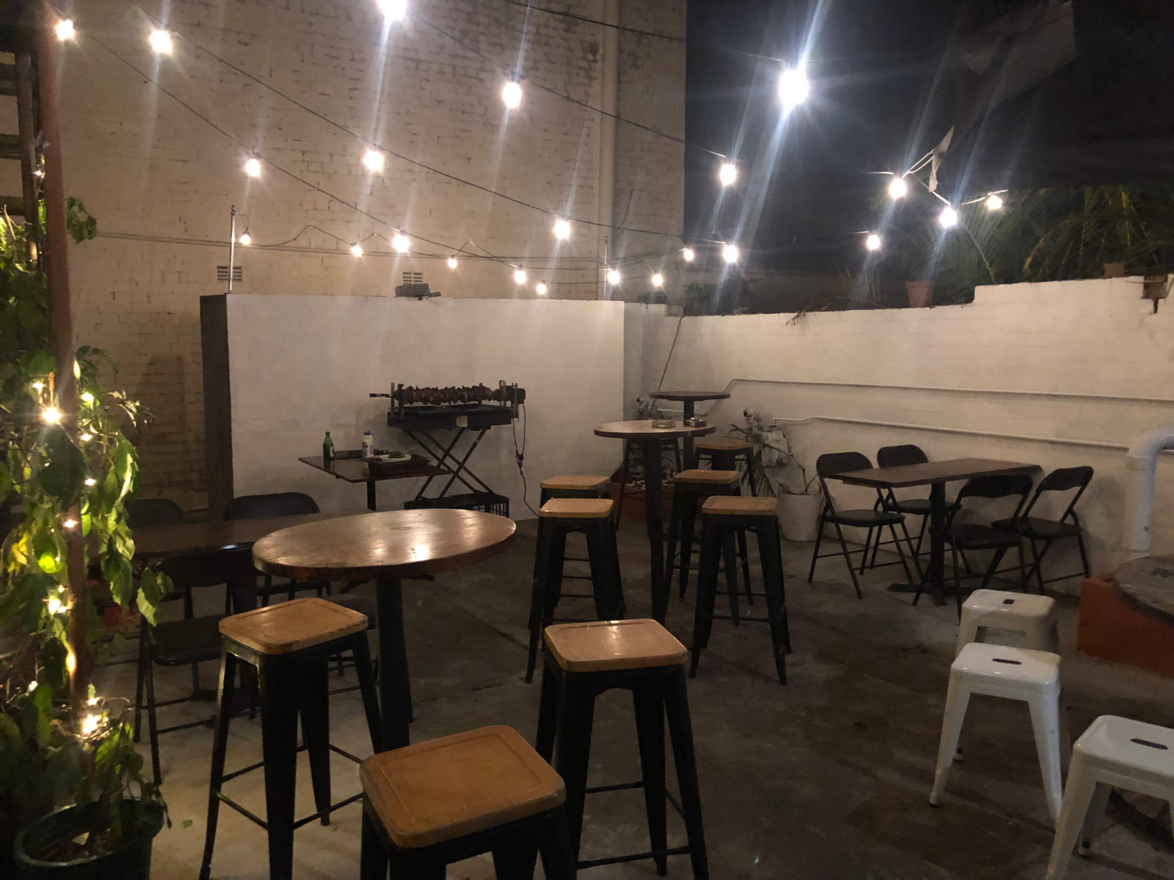 The image shows a dimly lit outdoor patio area with wooden tables and metal chairs, strung with overhead lights and a small grill in the background.