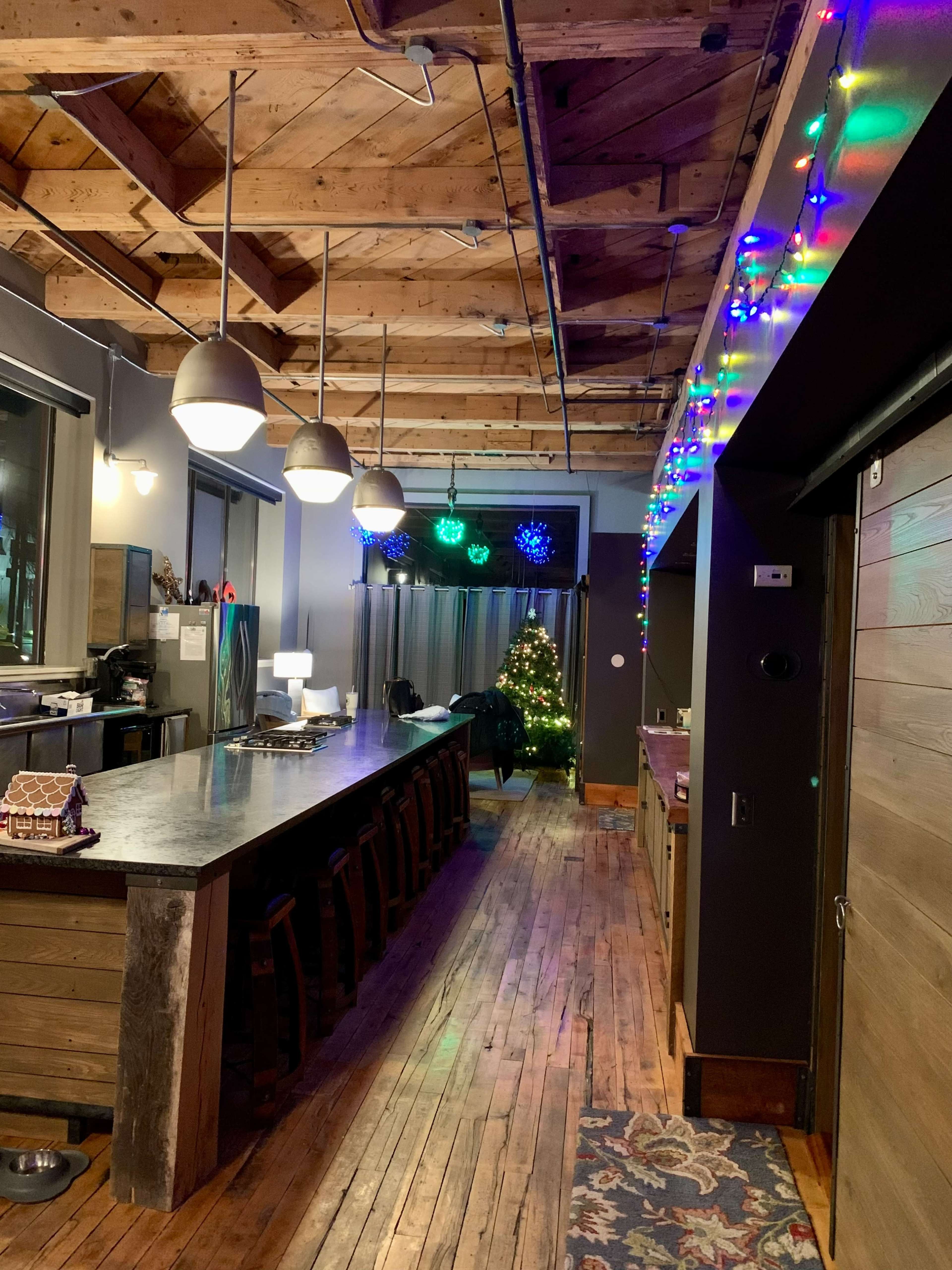 A spacious room featuring a long wooden bar with pendant lights, decorated with colorful holiday lights and a Christmas tree in the corner.