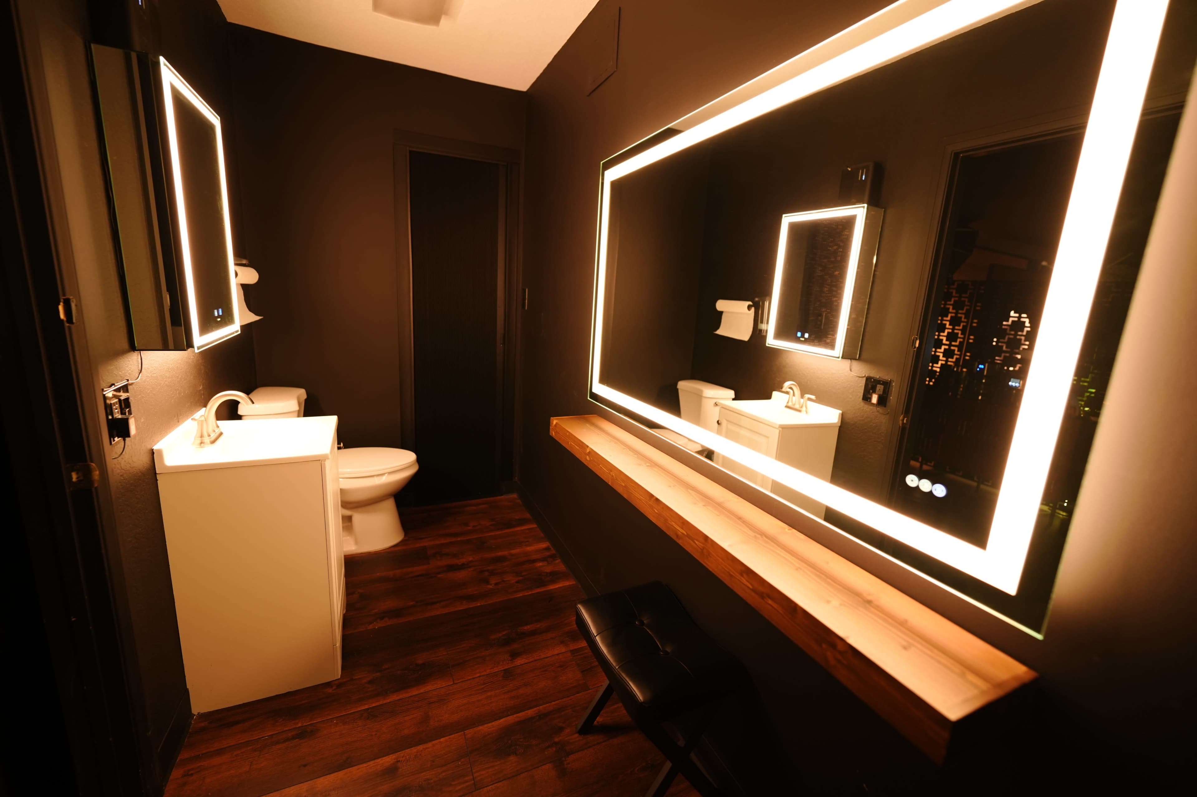 The image shows a modern bathroom featuring a white toilet, a sink area with a large backlit mirror, and a wooden shelf above a stool.