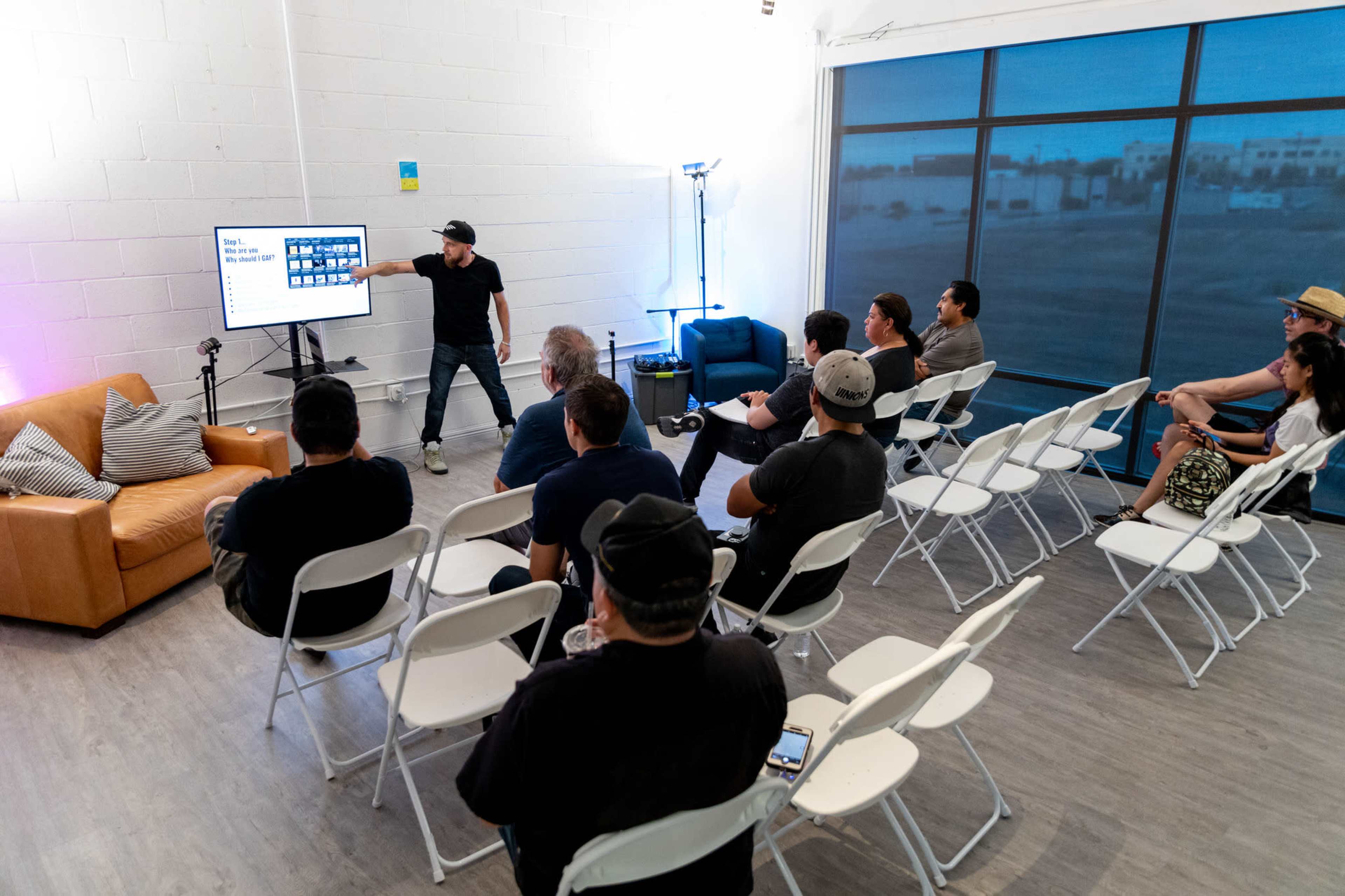 A presenter stands in front of a small audience seated in white chairs, pointing at a screen displaying various images in a well-lit room.