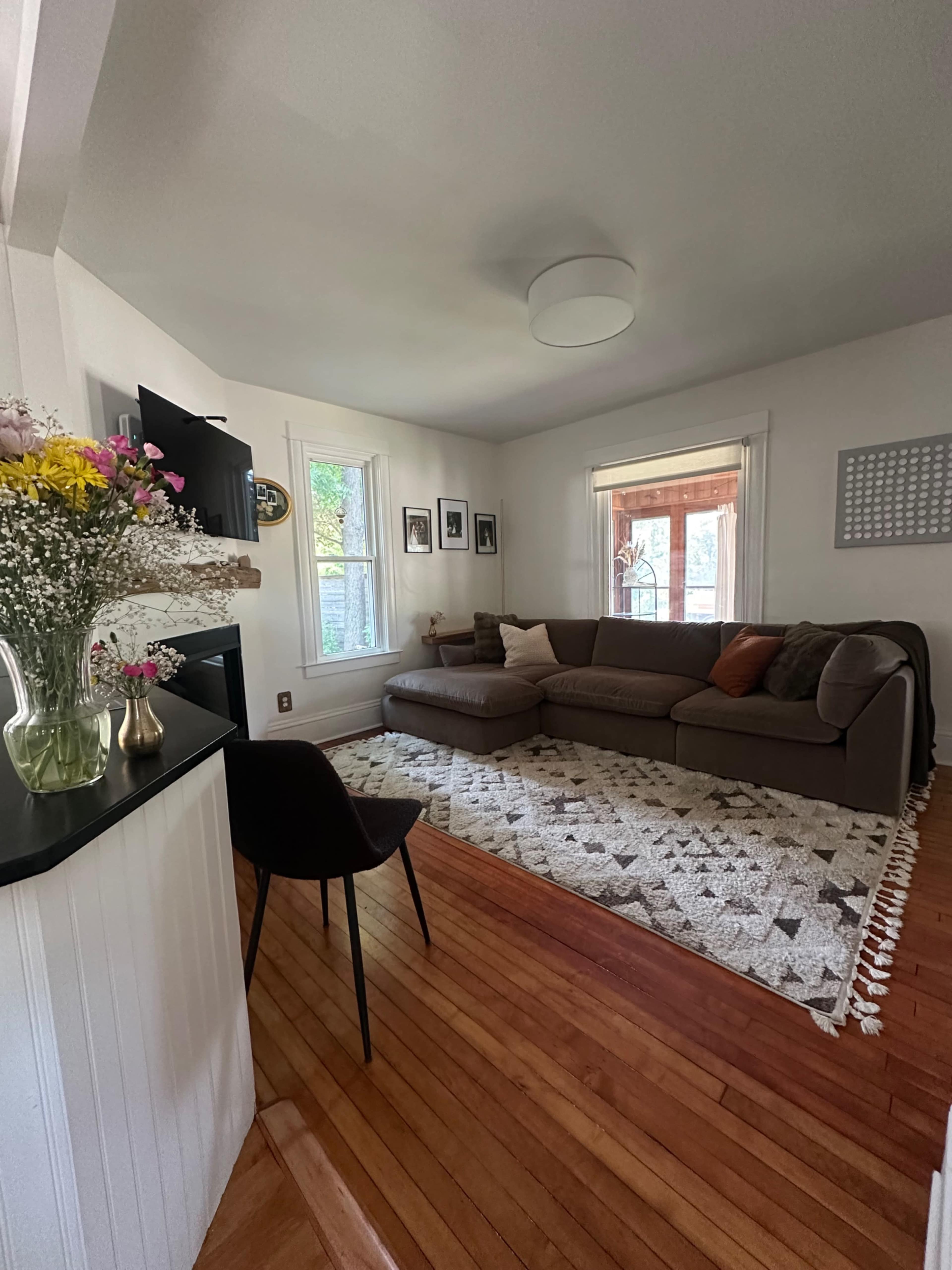 The image shows a living room with a large sectional sofa, a coffee table, and a patterned rug, accompanied by large windows and a flower arrangement on a side table.